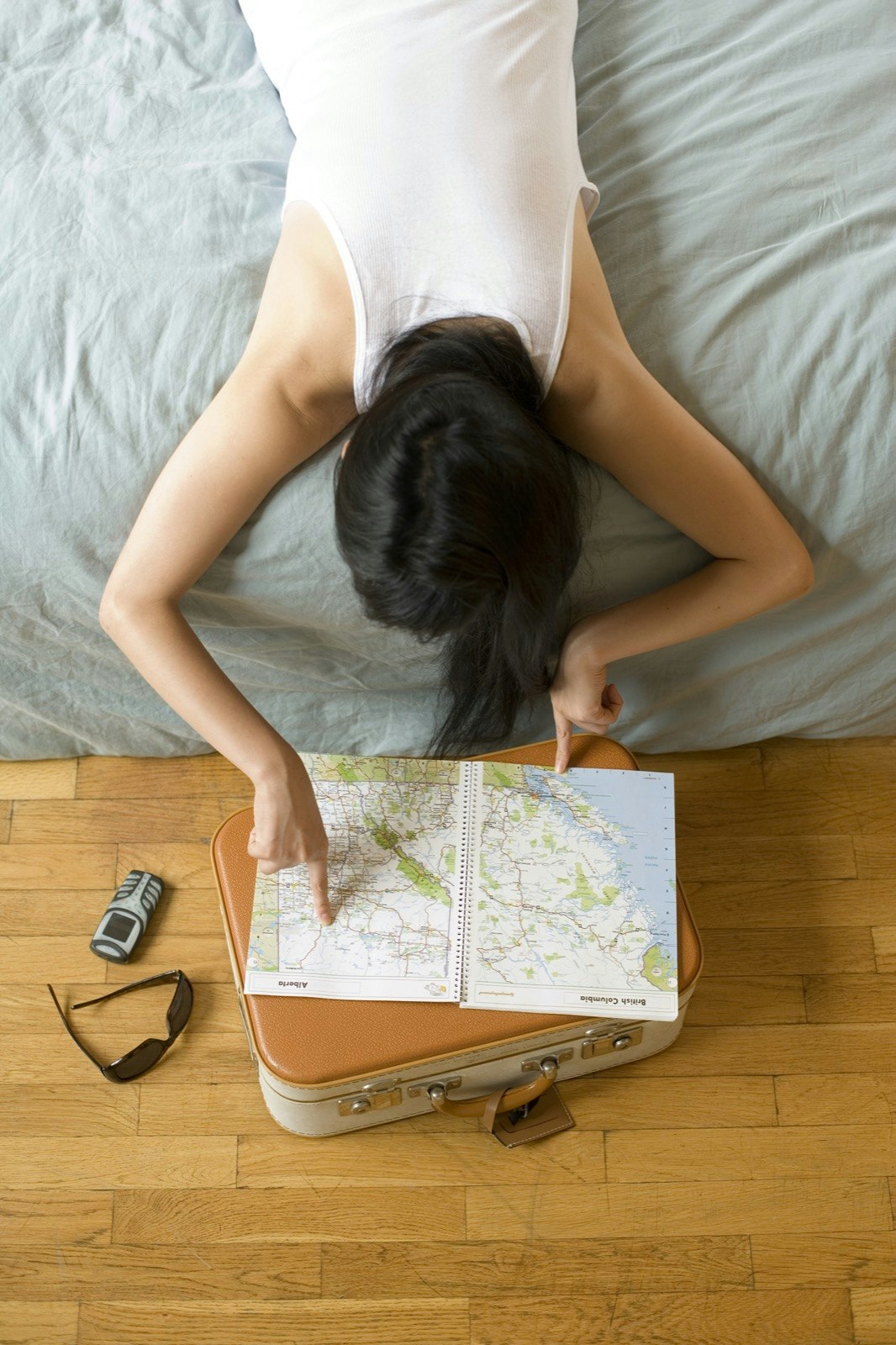 A woman lies on a bed and looks down at a map and suitcase