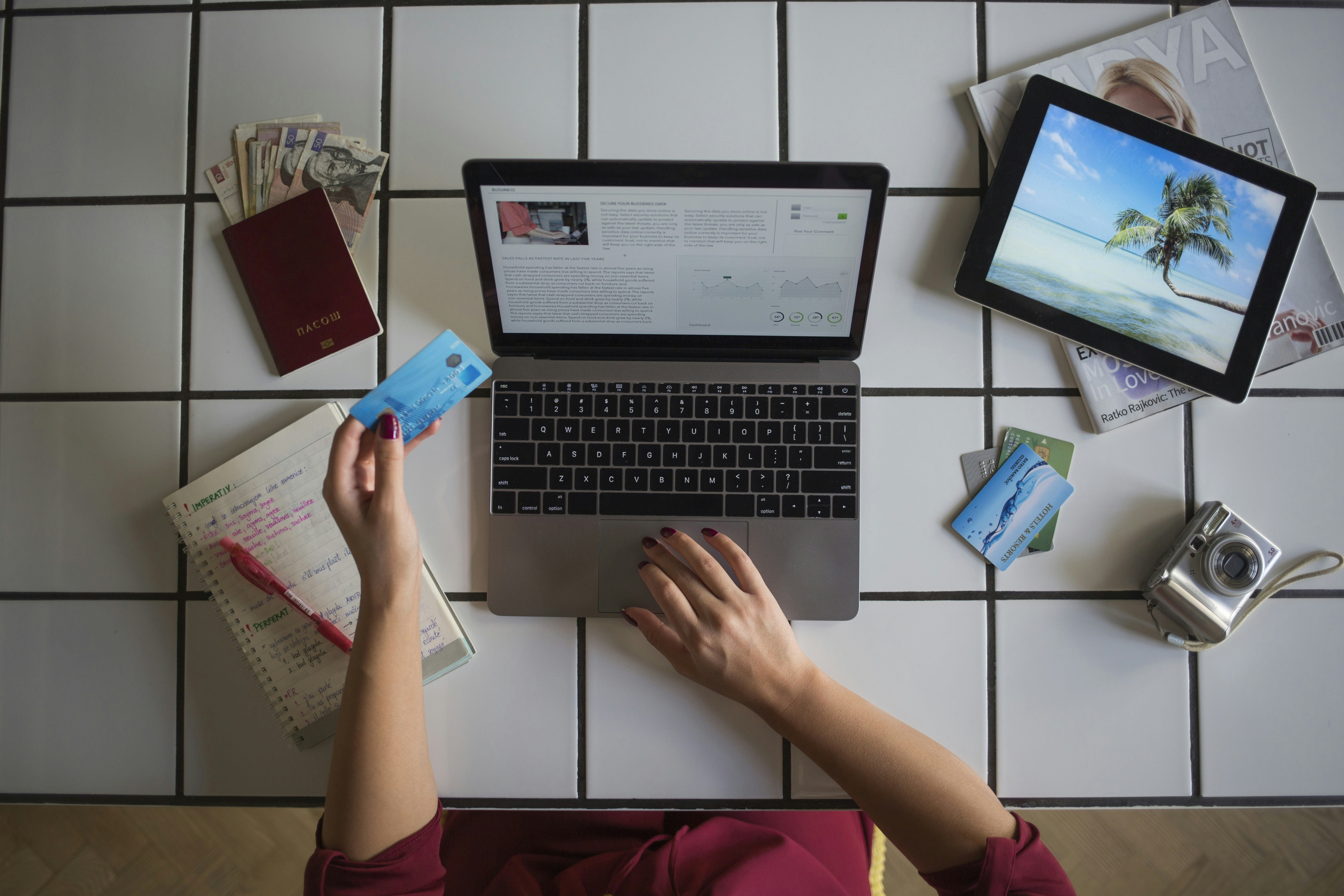 A top-down view of a person working at a laptop, planning their upcoming trip. Splayed out on the table around them is a passport, diary and foreign currency notes.