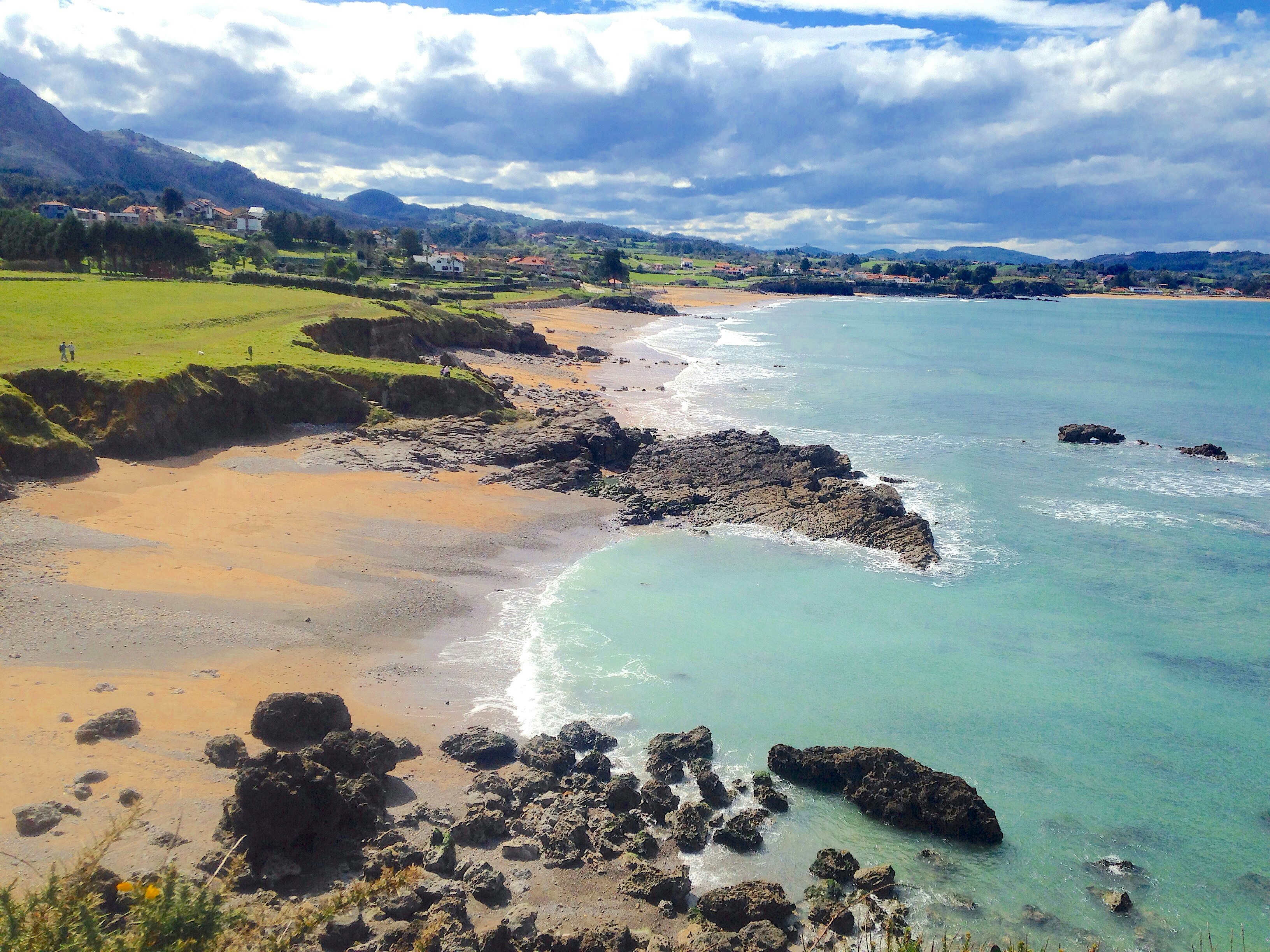 A coastal scene on the Camino del Norte; there's a rocky beach with calm turquoise water backed by pasture and rugged hills.