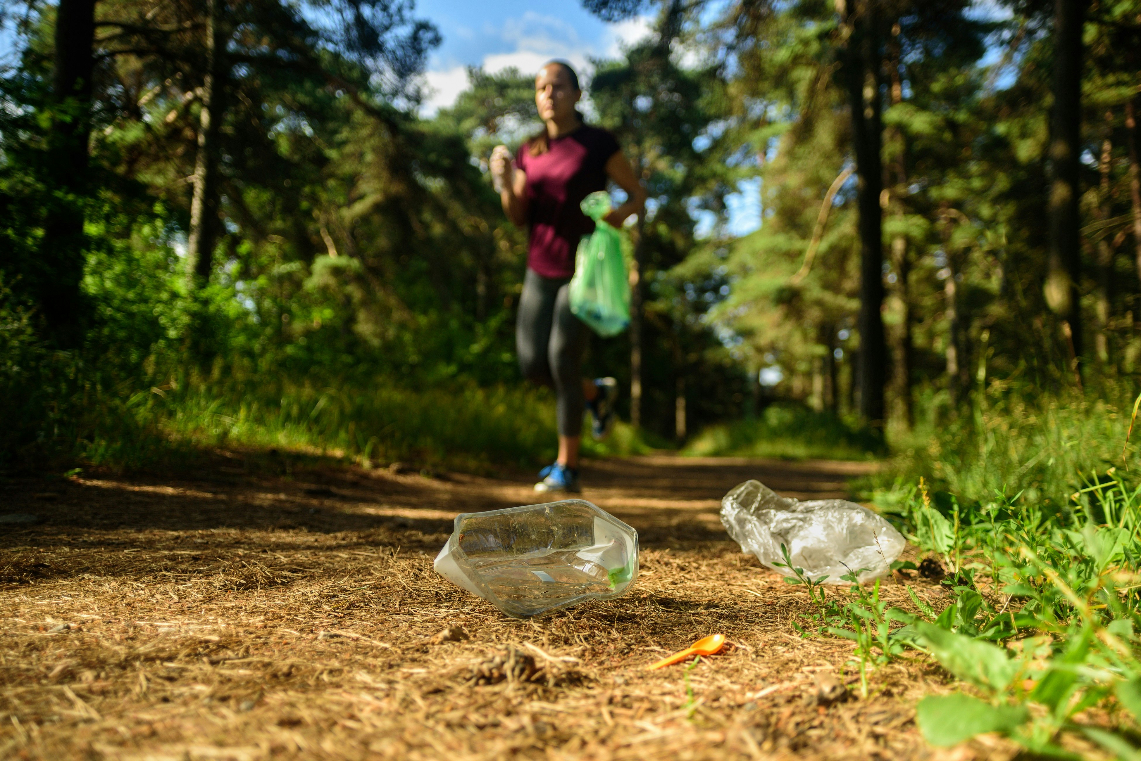 A woman in a bright green jacket and carrying a recycling bag jogs through a forest towards two discarded plastic cups in the foreground of the image.