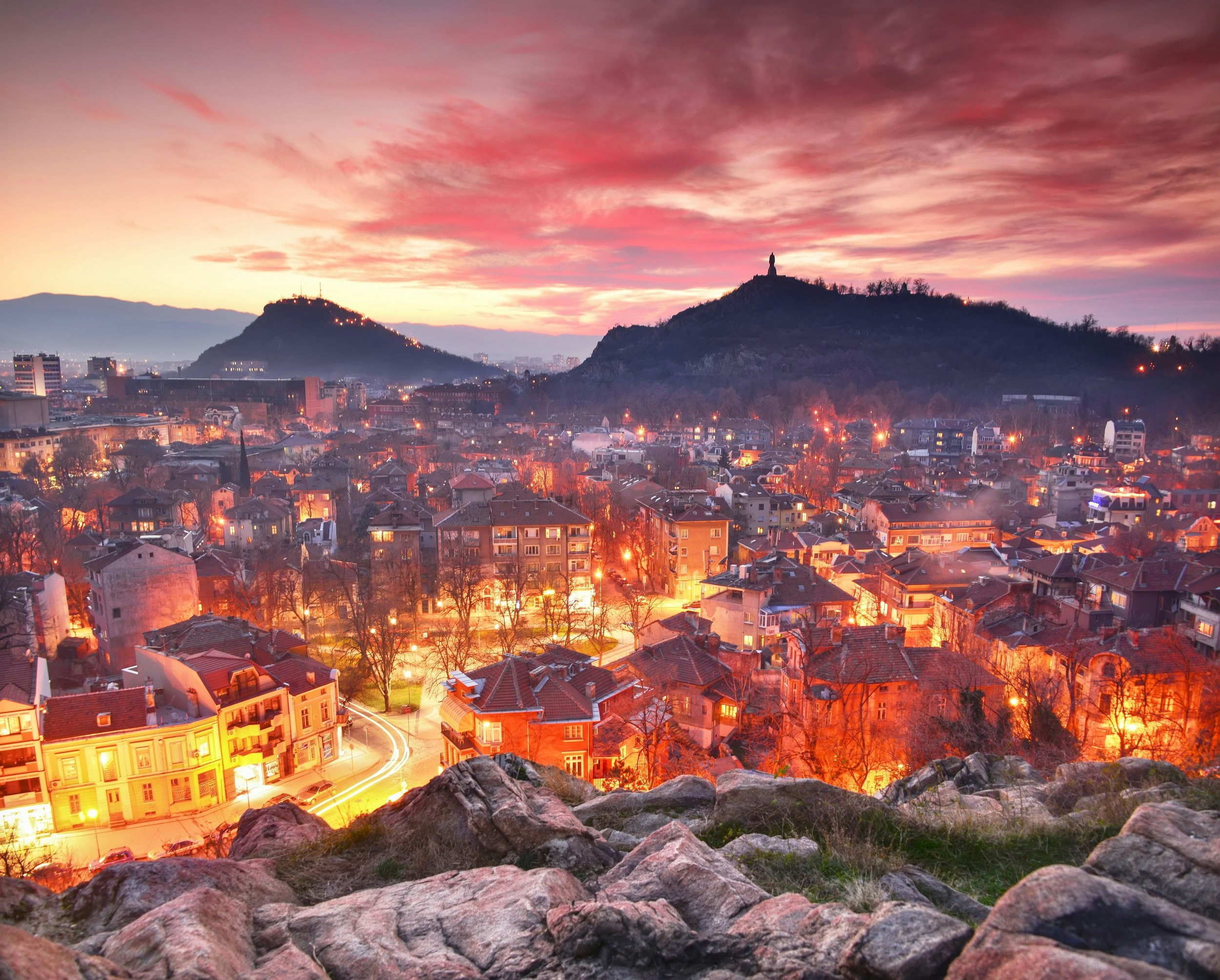 A cityscape with two hills in the background. The sun is setting, casting a pink hue across the sky