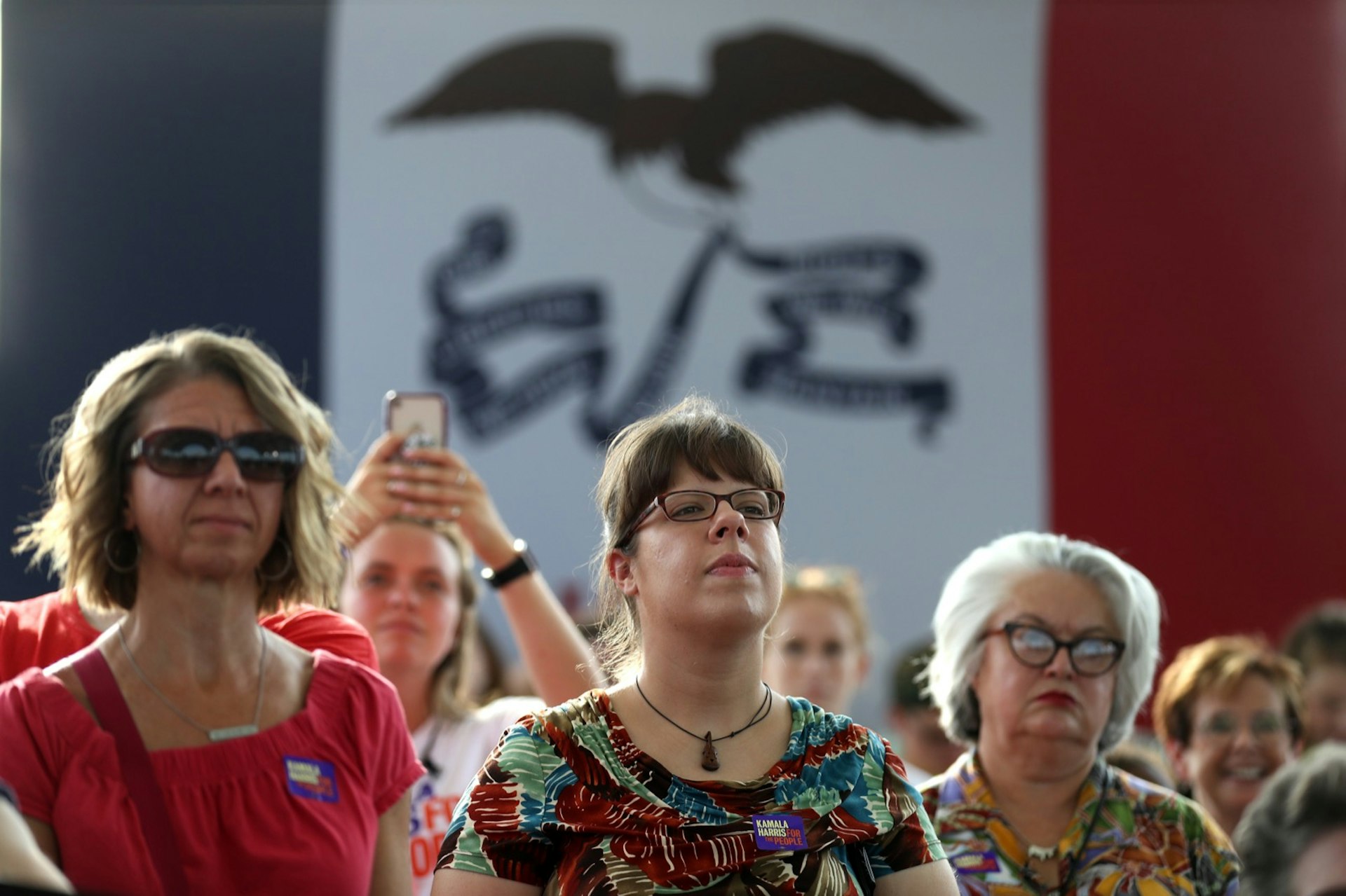 People listen to a candidate with an enormous Iowa flag behind them during the Iowa Caucuses in 2019; Things to do in Des Moines during the Iowa Caucuses