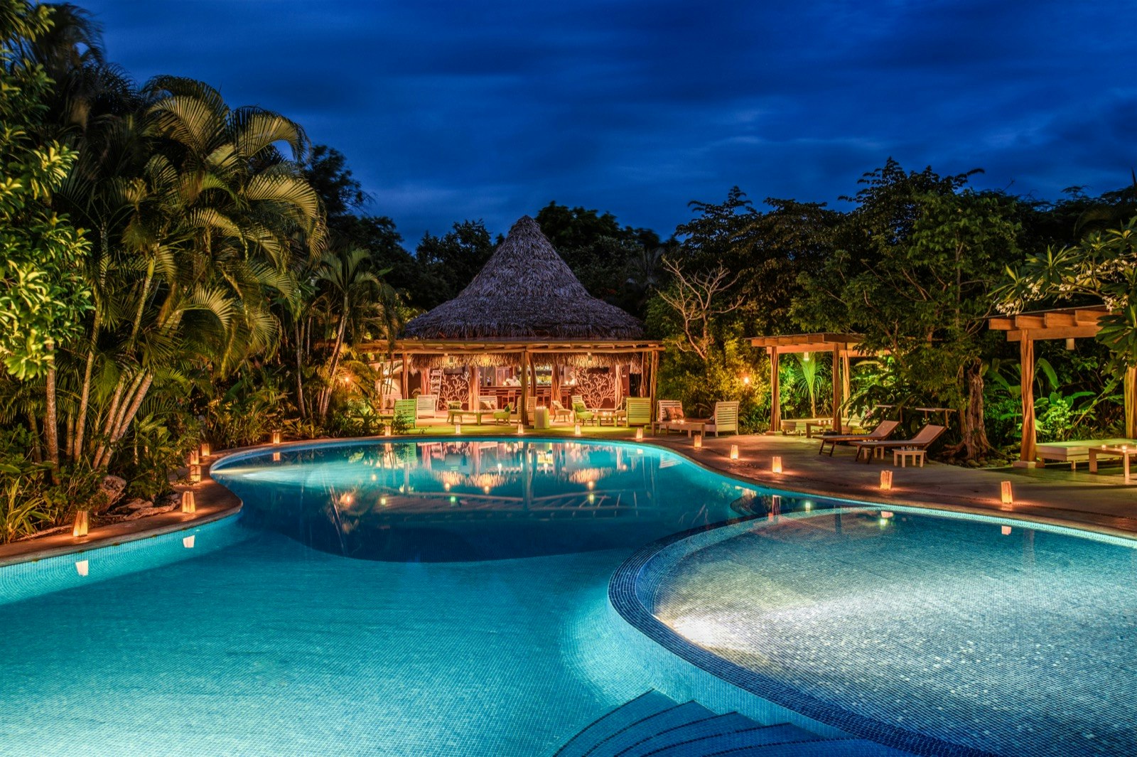 A large clear pool encircled by small lamps sits in front of a wooden pool-side restaurant; eco luxury resorts