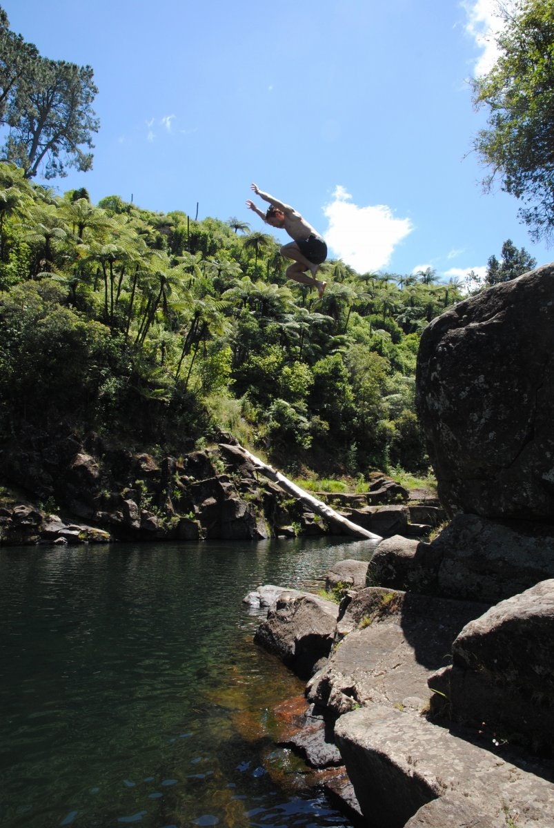 An image of a man water bombing into a small inland lake. It's a sunny day and the water is surrounded by rocks and verdant greenery. 