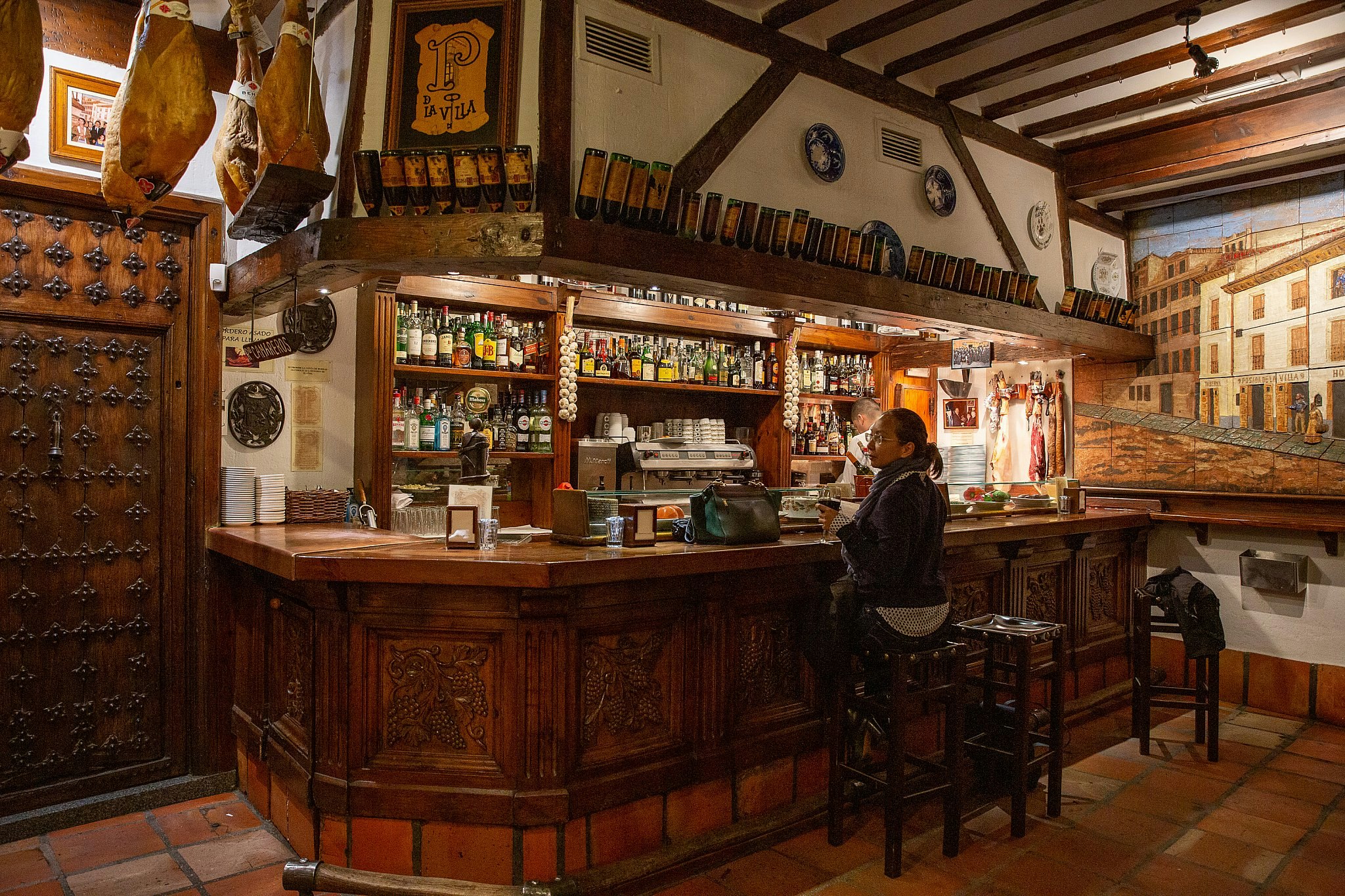 Posada de la Villa in Madrid: a lady sits at an intricately carved wooden bar; there is a mural on the far wall, and a beamed ceiling from which cuts of meat are hanging.