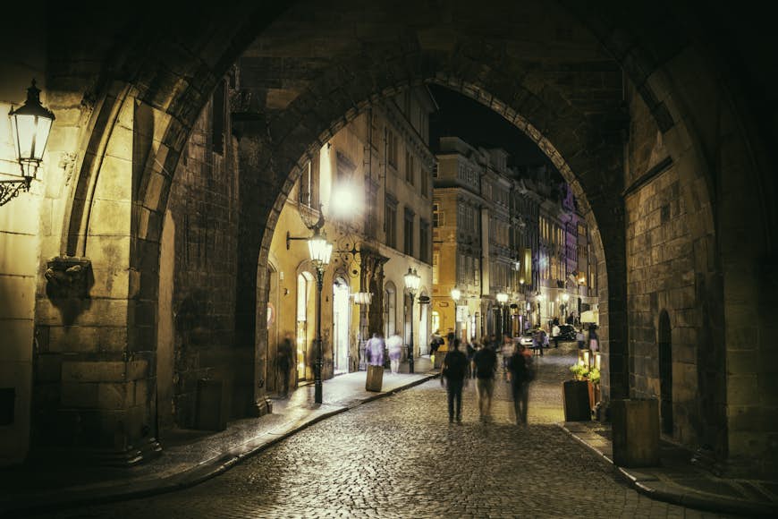 En grupp människor går nerför en kullerstensgata mot en livlig gata i Prag på natten