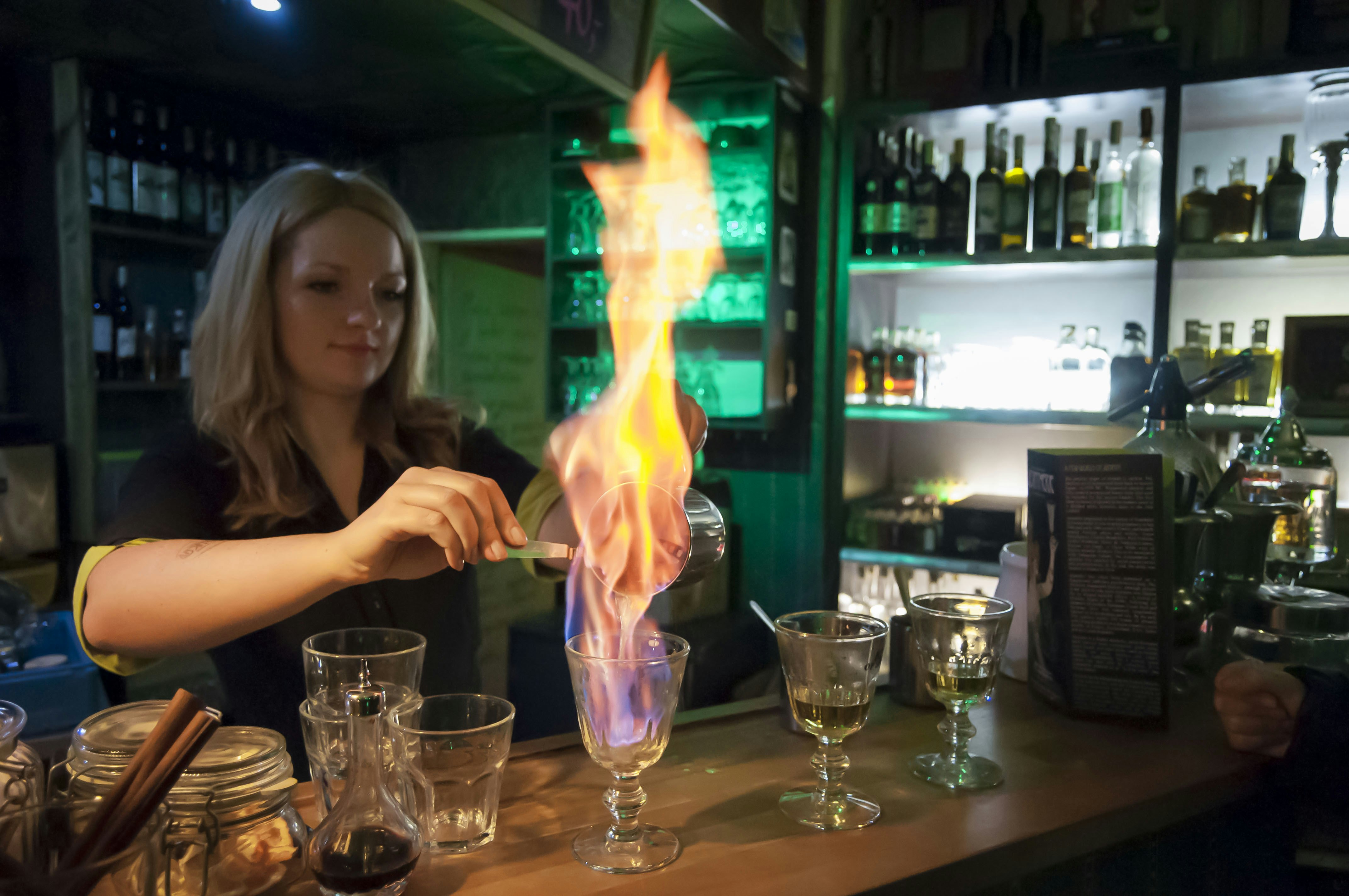 A fairly large flame shoots from a glass as a bartender pours absinthe into it. 