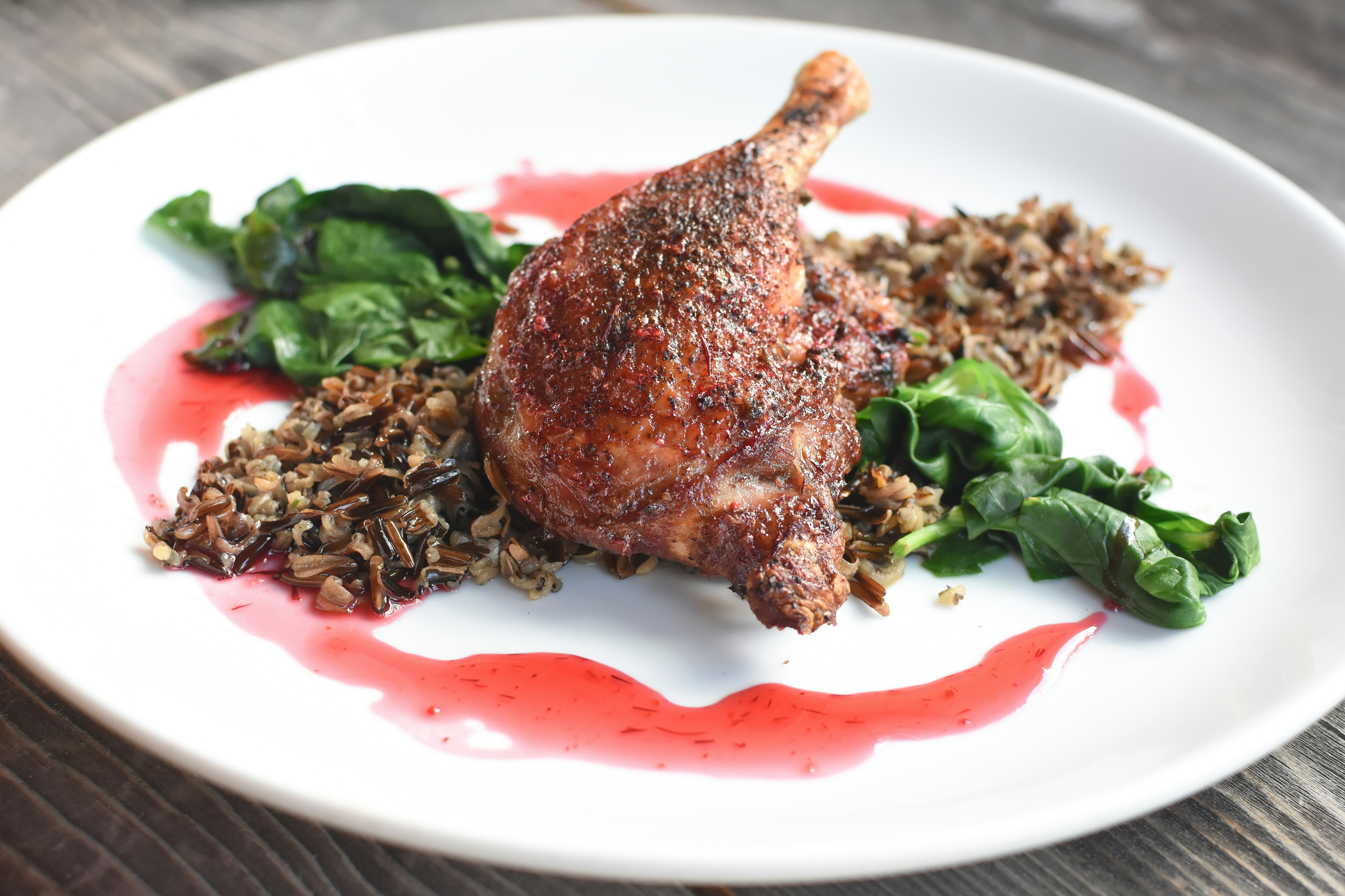 A piece of roasted duck lies on a bed of wild rice and greens on a white plate