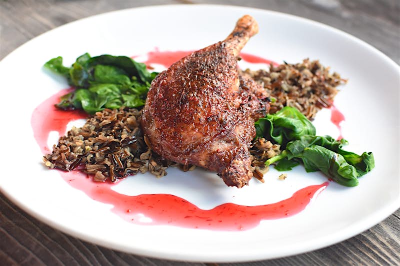 A piece of roasted duck lies on a bed of wild rice and greens on a white plate; Indigenous food