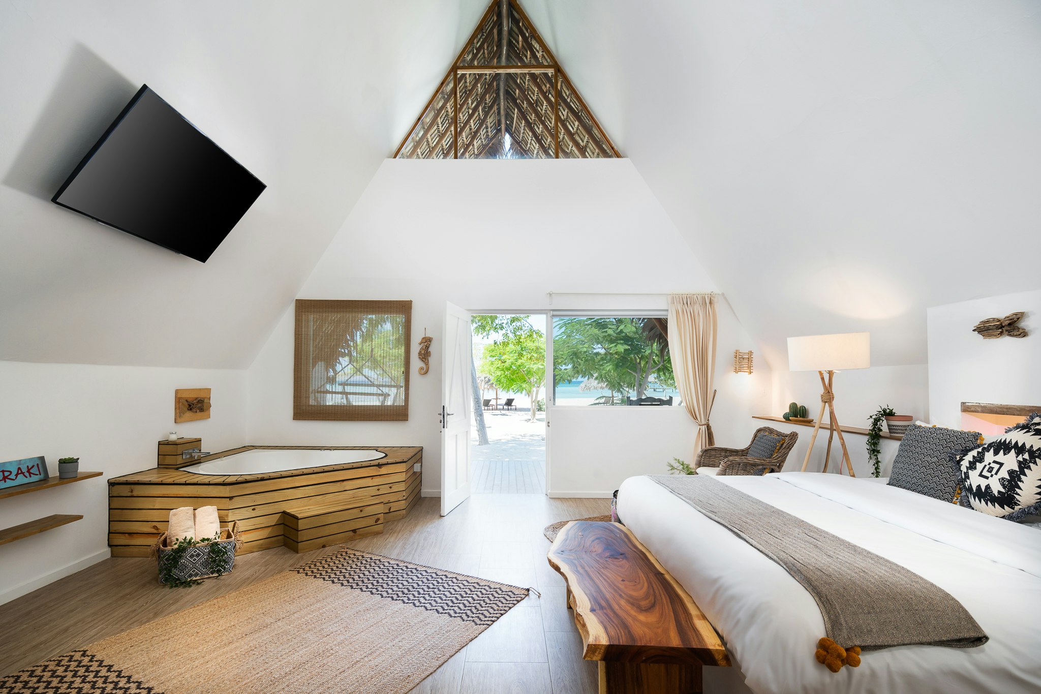 The pristine white interior of a hotel room at Punta Rucia lodge. There is a wooden bathtub to the upper left side of the room, a large flat-screen TV high on the wall, a woven mat and a well-made bed. The door of the room is open showcasing a view of trees along the beach.