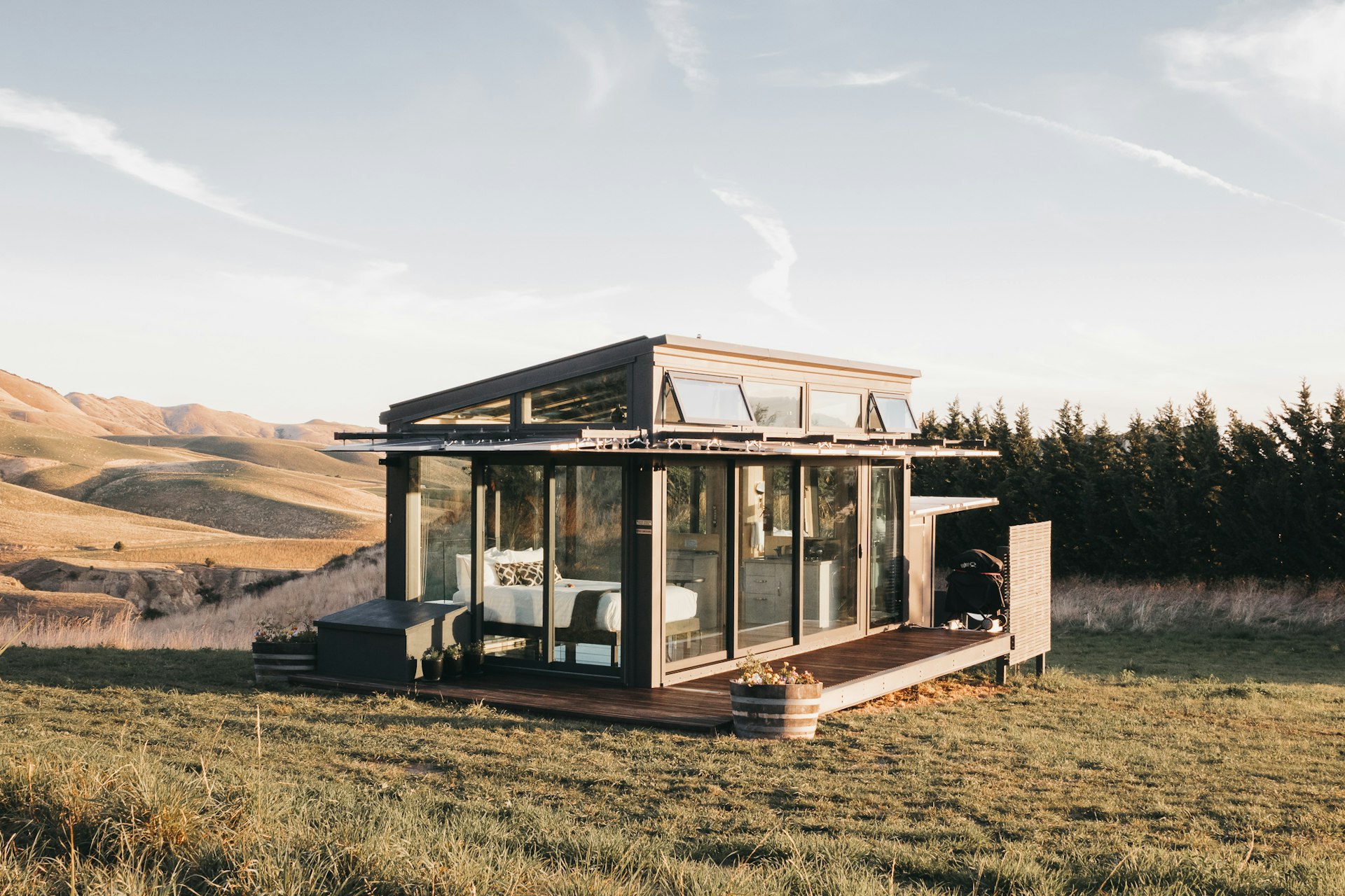A simple wood-and-glass chalet stands on grass with a hedgerow behind it. Beyond, there are barren, rolling hills.