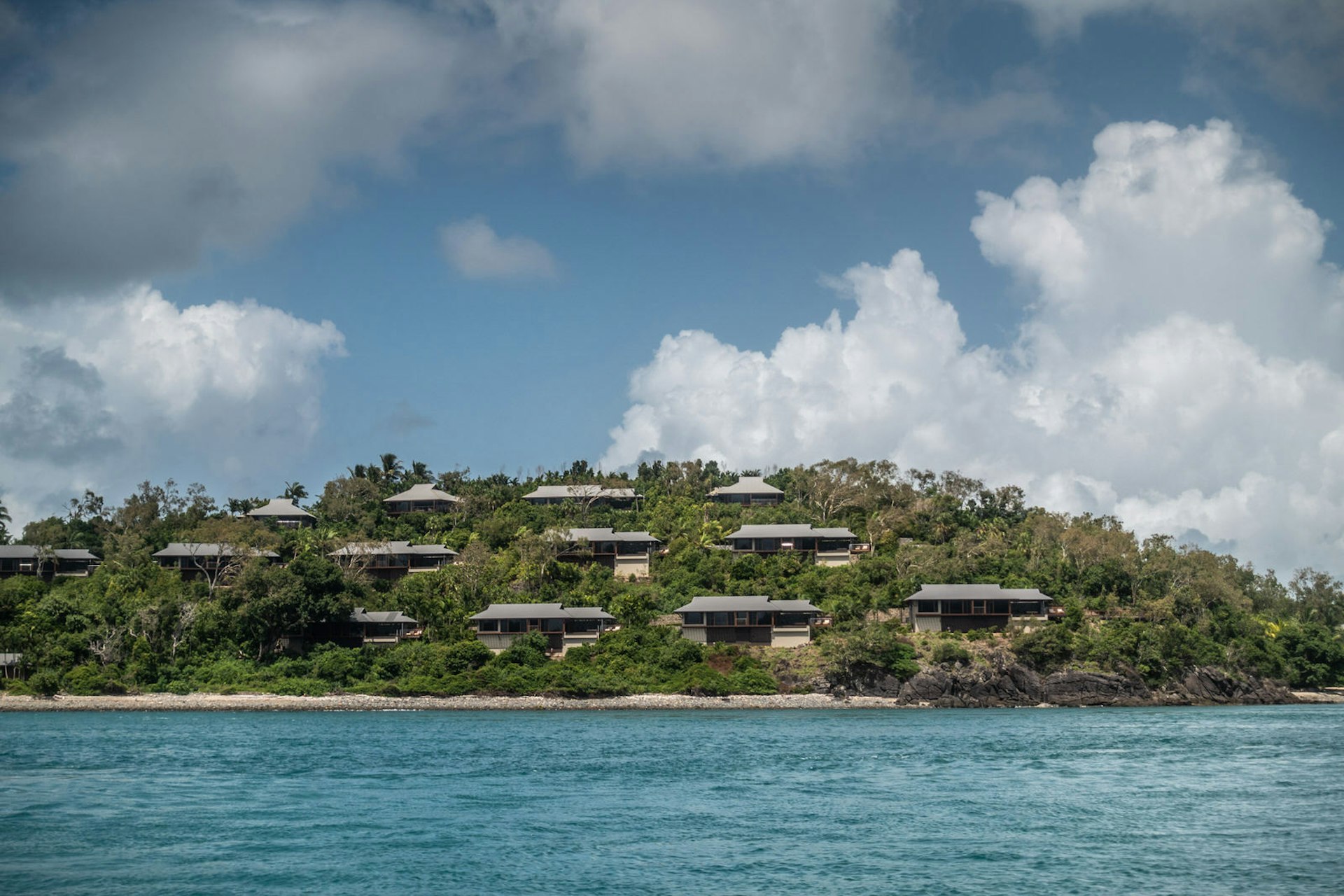 Villas at the upscale Qualia Beach Resort with a private beach, built on a green hill overlooking the coral sea. There's a blue sky with white cloudscape.