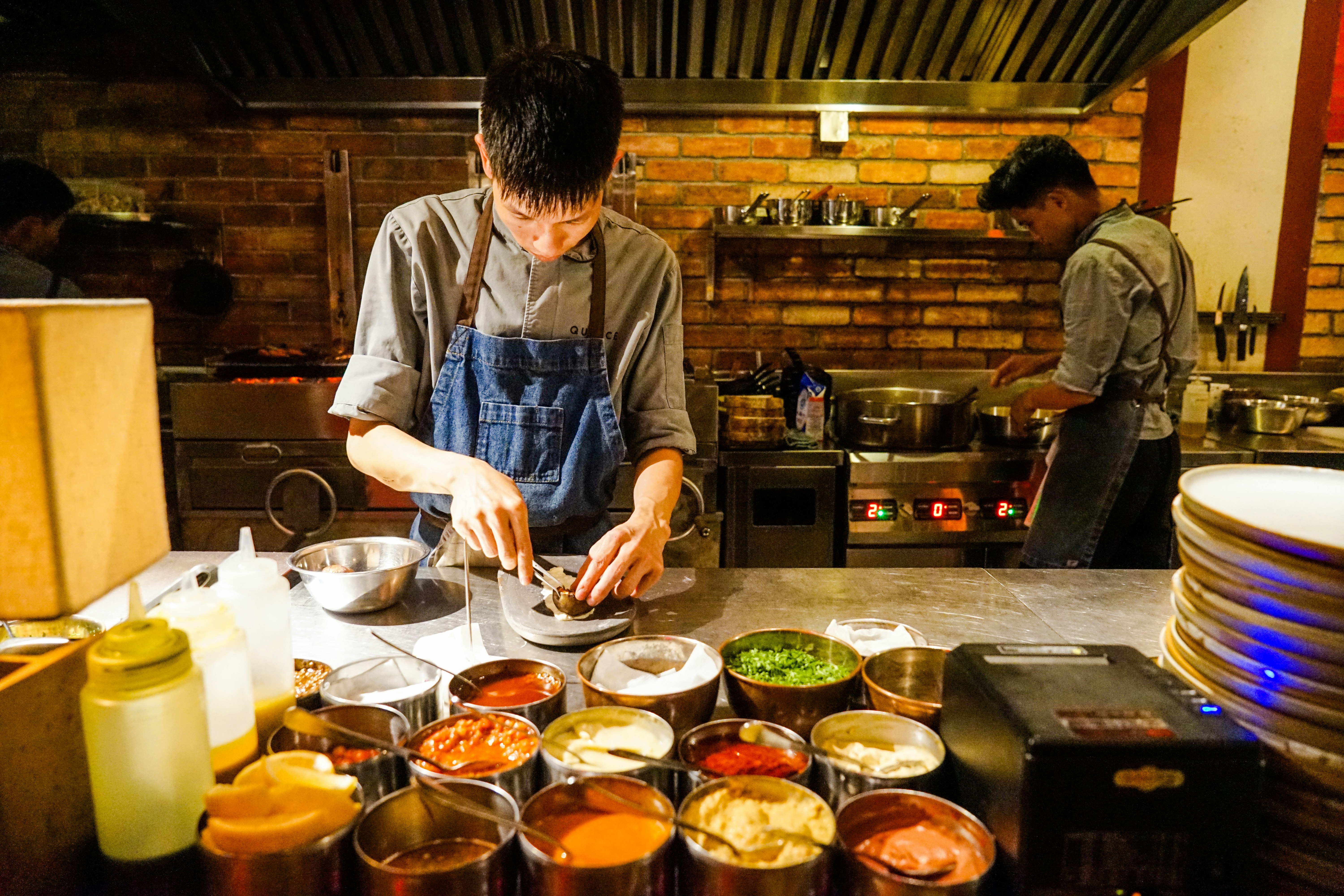 A chef plates a dish in a kitchen at Quince Eatery in Ho Chin Minh City. In front of the cook is a tray filled with an assortment of sauces. In the background, a man works over a pan.