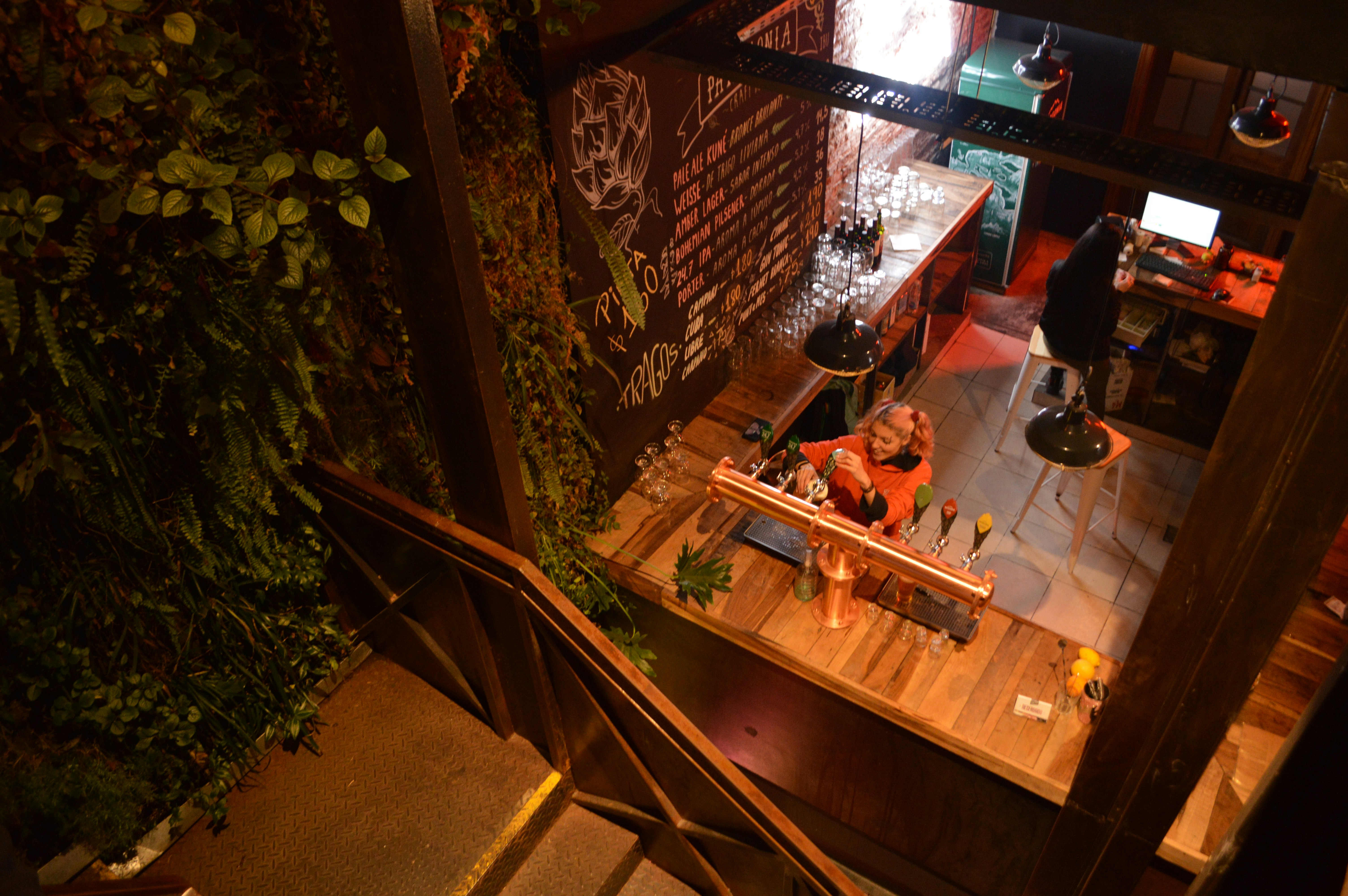 Overhead view of a bartender filling a glass with beer on tap. Behind the bartender and woman sits on a bar stool facing a computer screen; Buenos Aires rooftop bars