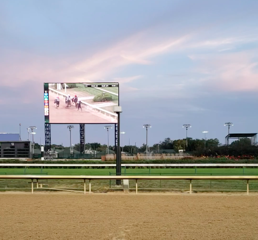 A side view of the tan dirt track at Churchill downs with green grass in the background and a blue sky with wispy white clouds. Over the track is a large jumbotron viewing screen that shows horses and jockeys actively racing
