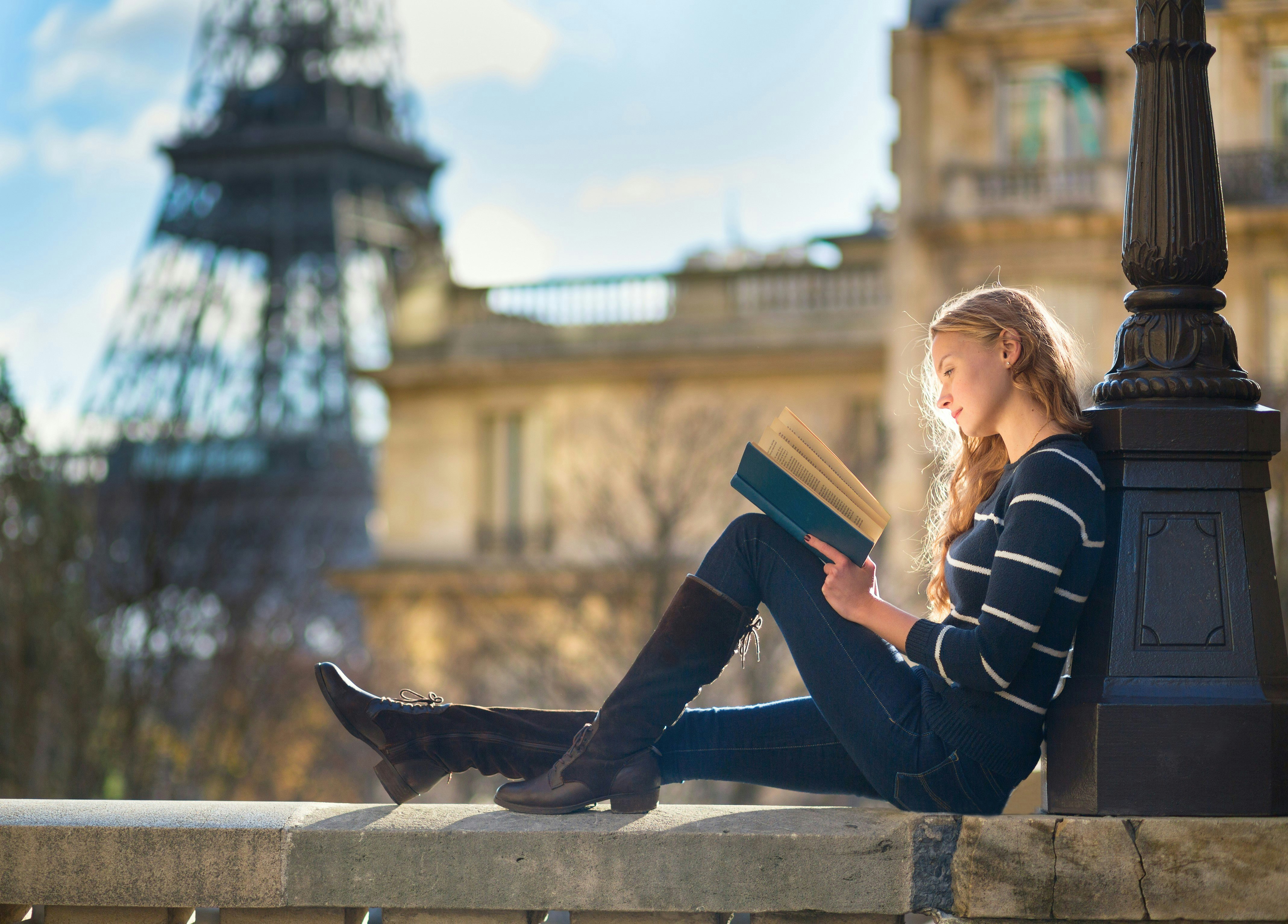 A woman sits on a wall in Paris reading a book. In the background the Eiffel Tower is visible. 