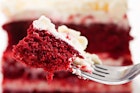 Extreme closeup of a bite of red velvet cake on a silver fork. In the background you can see the entire cake