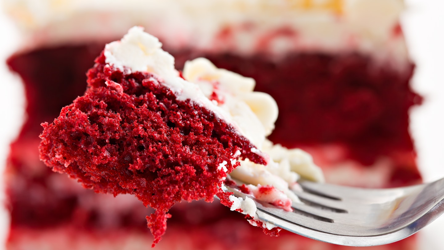 Extreme closeup of a bite of red velvet cake on a silver fork. In the background you can see the entire cake