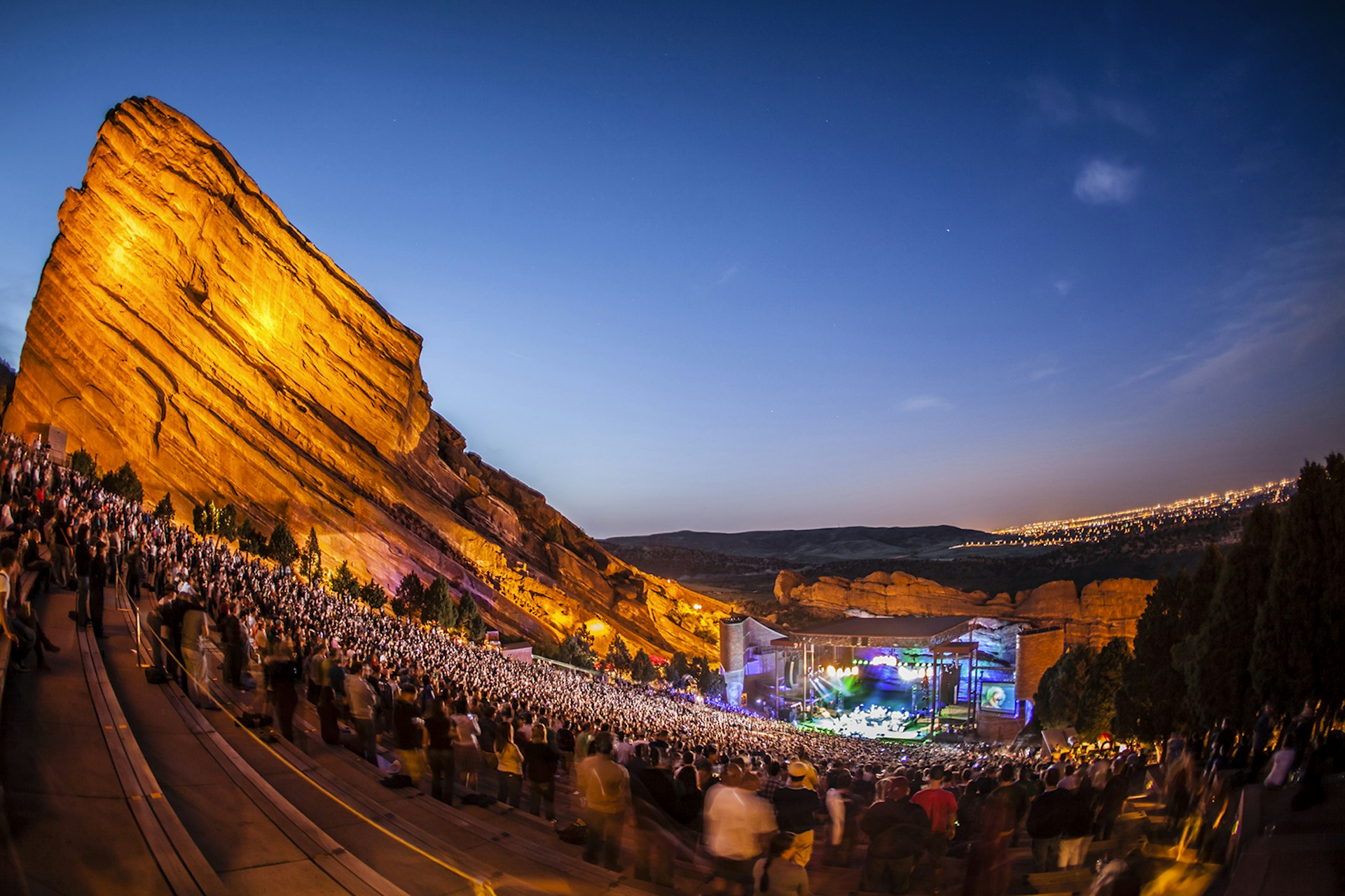 A crowd attends an evening concert at Red Rocks Amphitheater; one of the stone walls is visible, and the city of Denver glows behind the stage. Colorado.