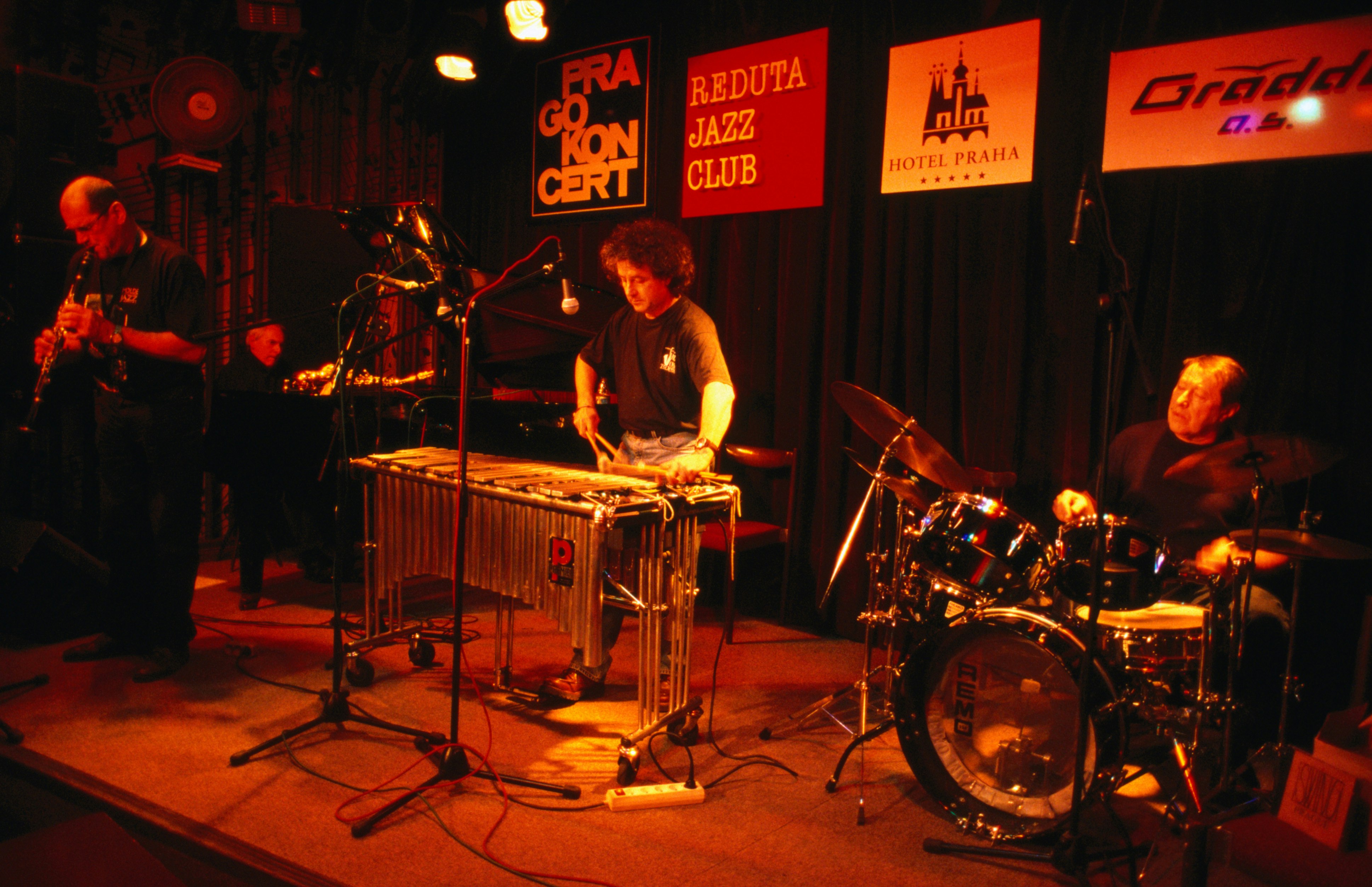 A jazz quartet made up of a drummer, piano player, clarinetist and a man on a xylophone play on a dimly lit stage at the Reduta Jazz Club