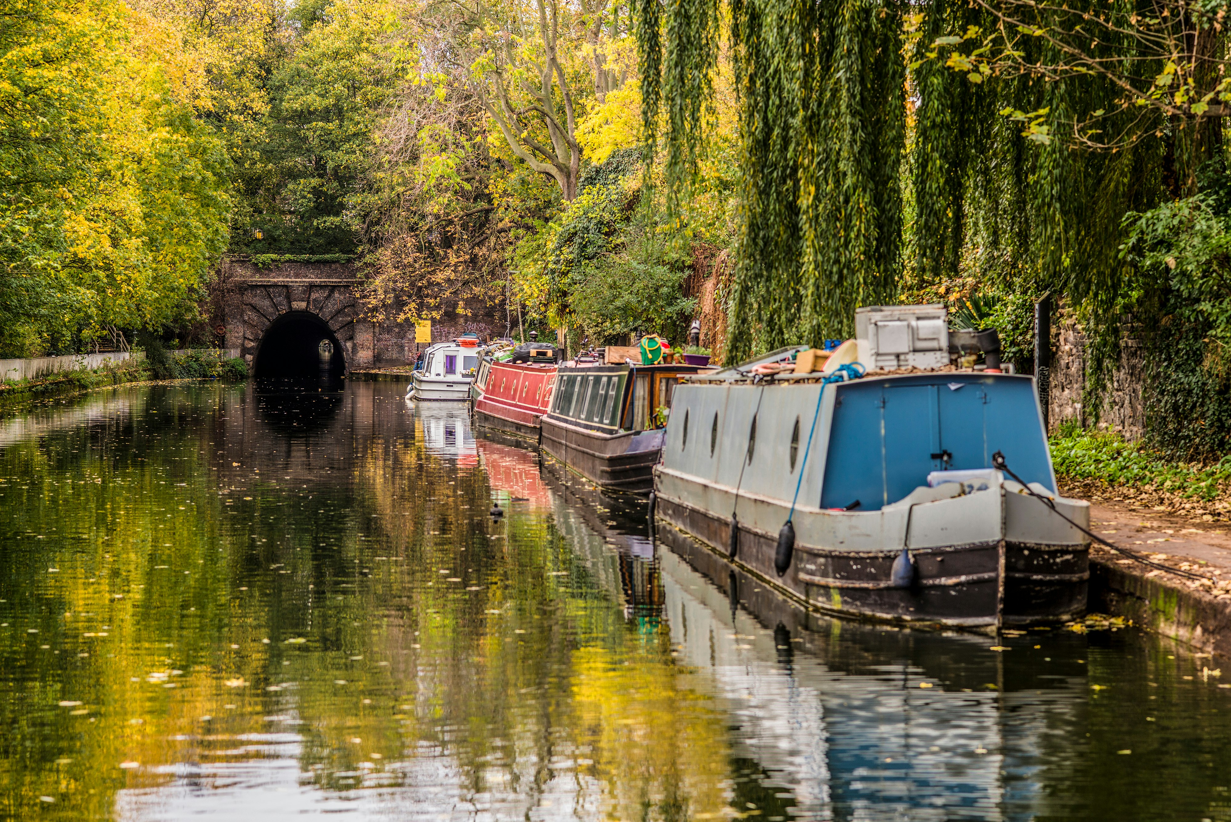 Canal boats docked alongside a narrow canal, with fall colors on the banks and a tunnel in the background