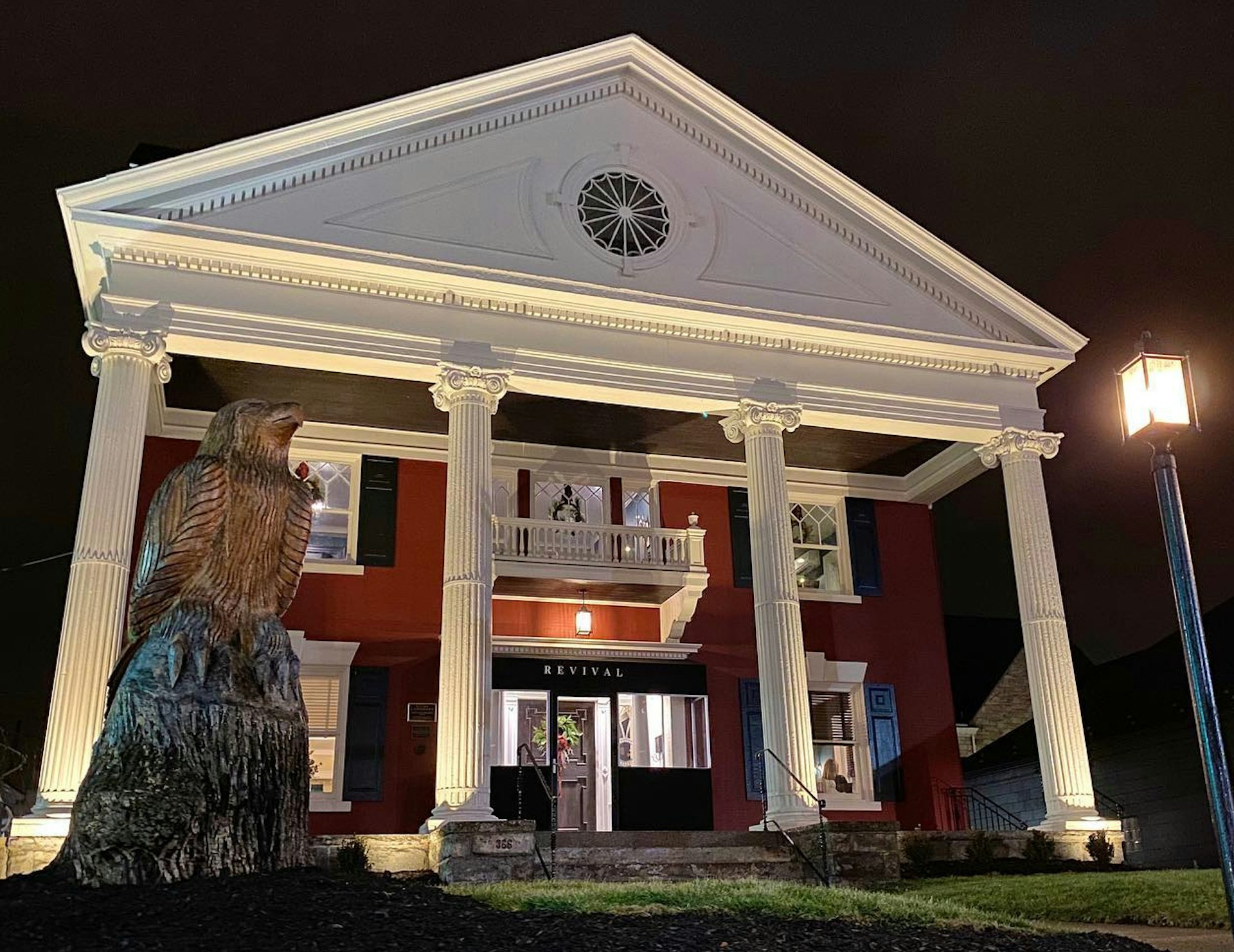 The exterior of Revival on Lincoln at night shows a red brick two story building with four white Ionic columns and a Federal style roof with a large sculpture of an eagle in the foreground