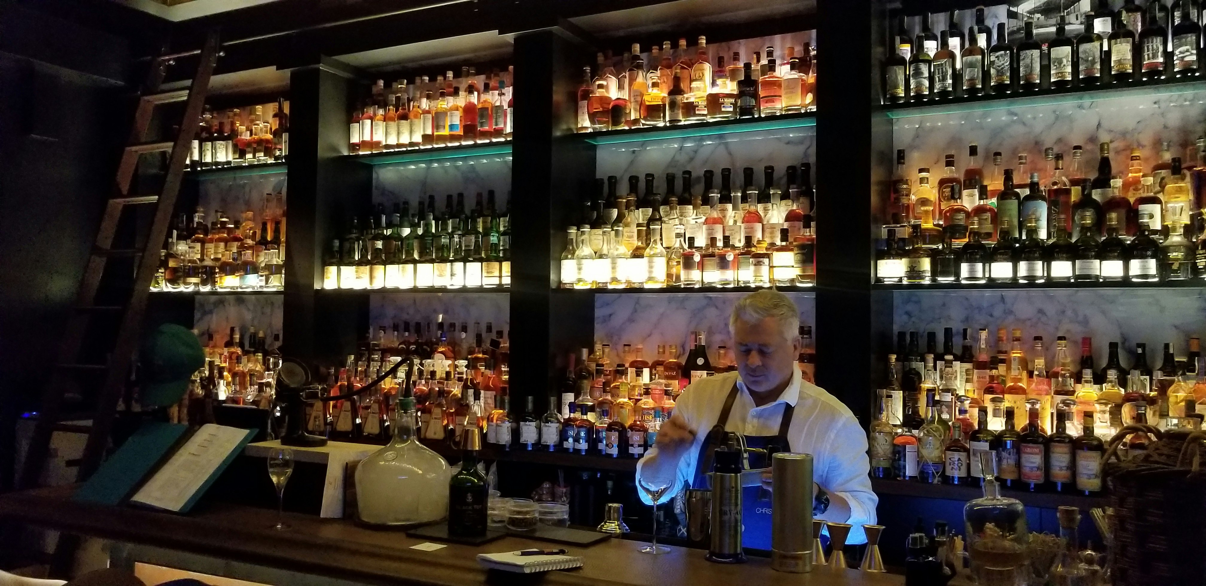A bartender wearing a dark blue apron mixes a drink in front of a large rum display 