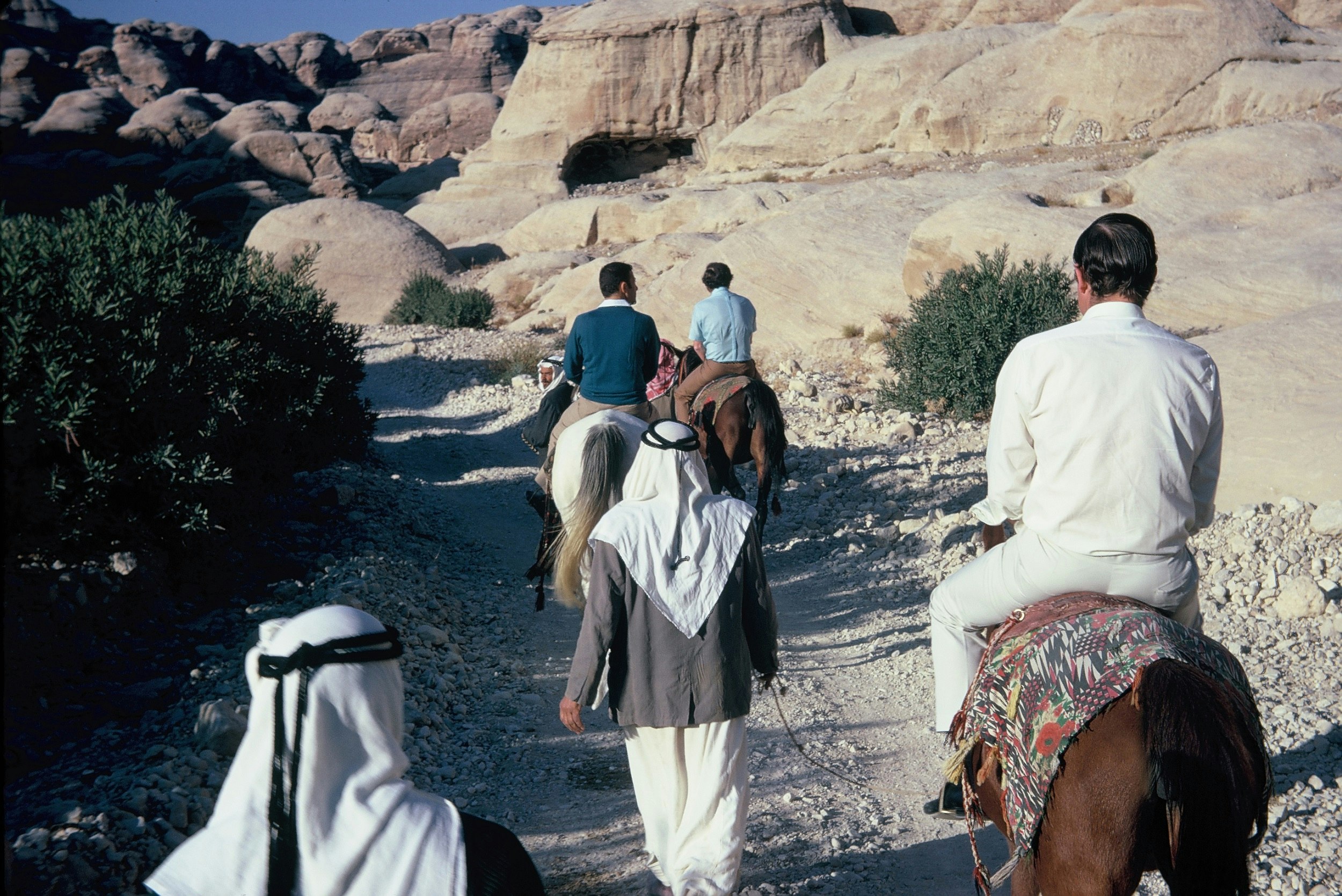 Three men on horseback are led by walking guides wearing Middle Eastern headdress; they are on a stone path leading through rocky mountains.