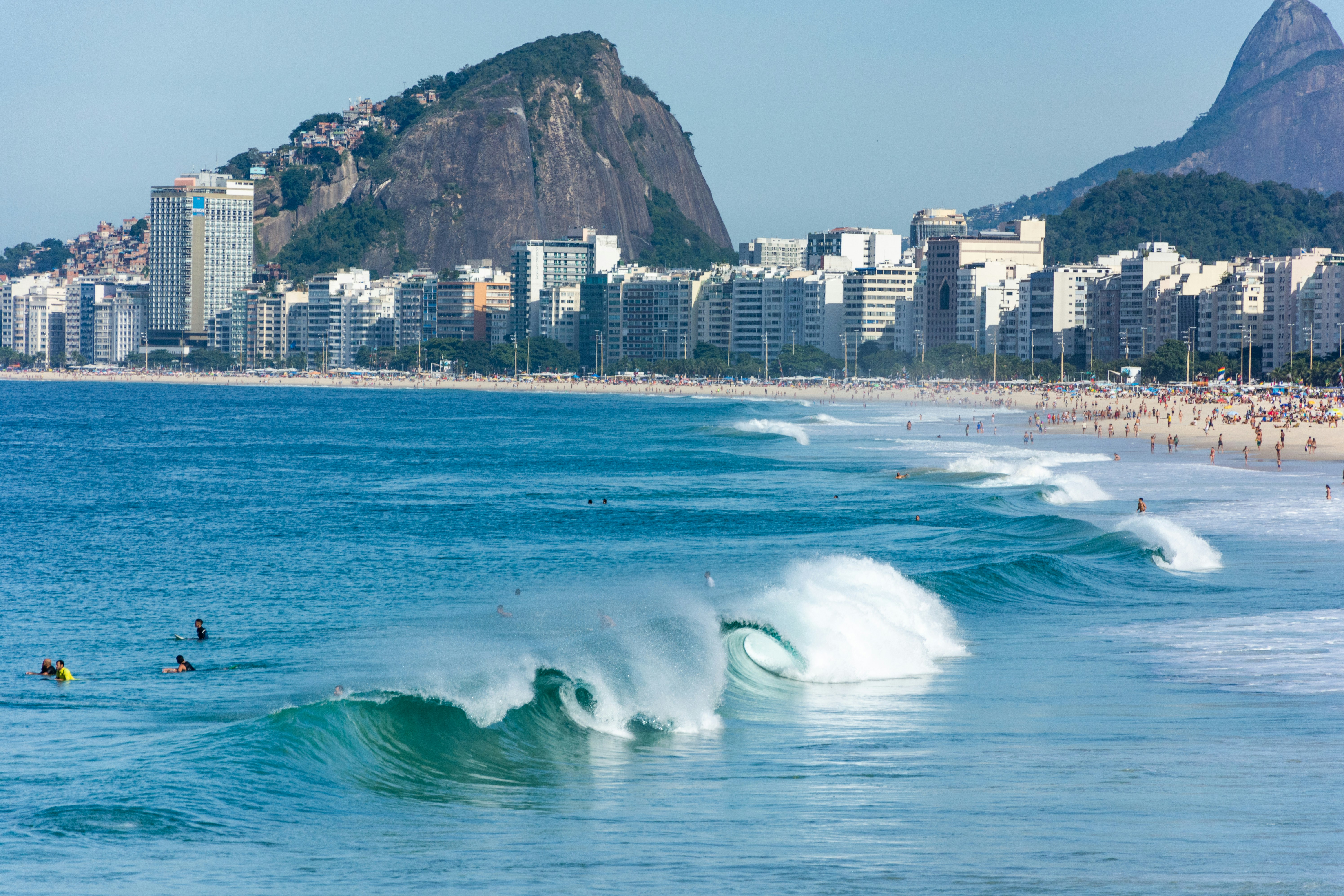 Waves crash to the shore on the busy Copacabana beach, in Rio de Janeiro, on a sunny day. People are swimming in the ocean.