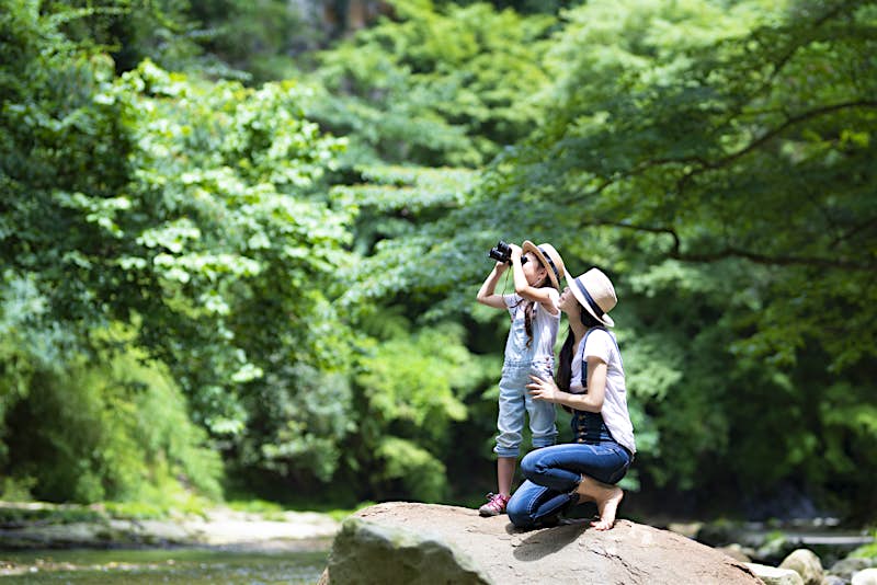 A mother and her daughter, both in denim overalls, explore a river while standing on rocks. The daughter is looking into the green woods with binoculars; kids outdoor adventures