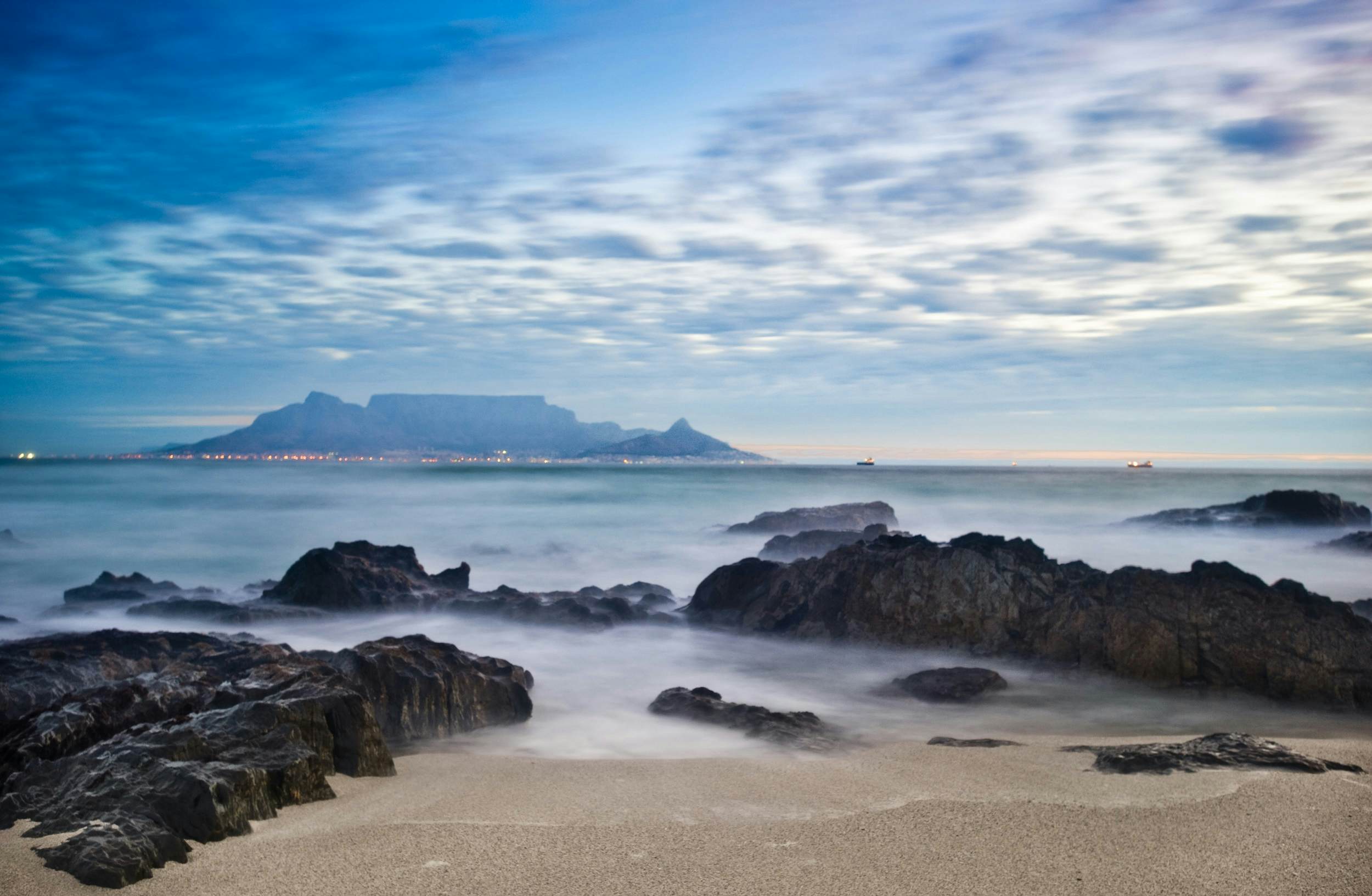 Looking across sands trapped between rocky outcrops on the beach of Robben Island; across the waters in the distance is Cape Town and the unmistakable flat-topped Table Mountain.