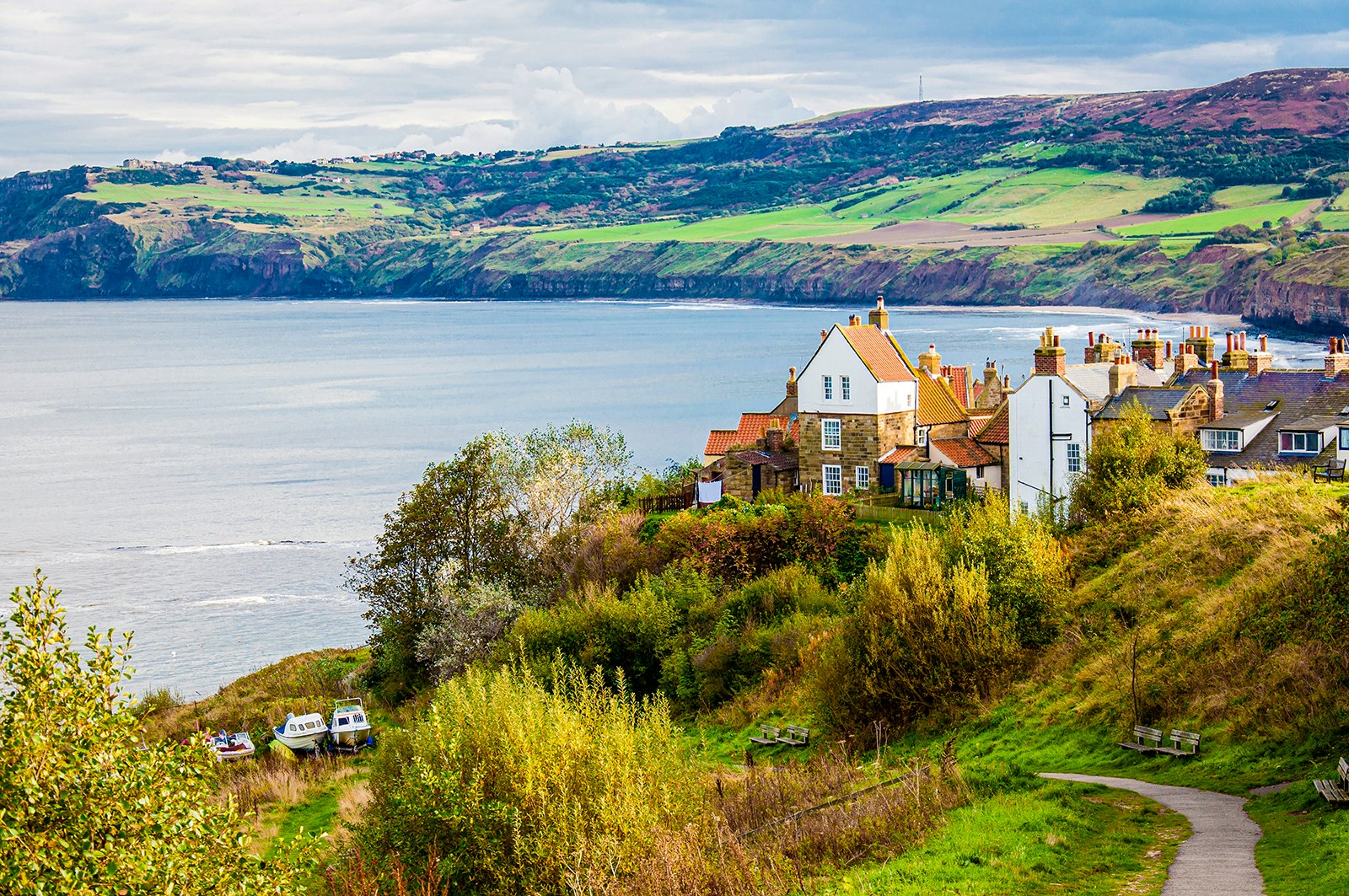 A small path leads to a fishing community with historic white buildings with orange roofs. The town sits on the edge of a bay with rolling green hills in the background. England.
