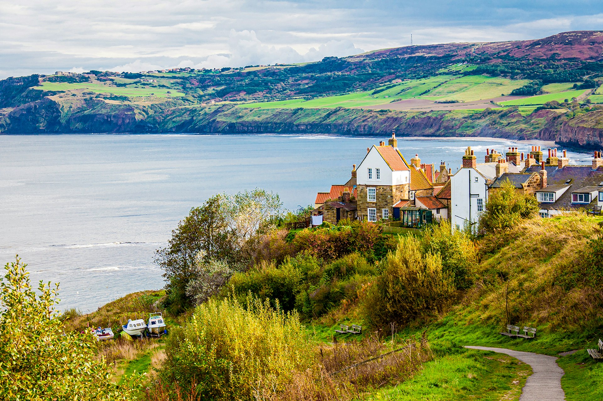 A small path leads to a fishing community with historic white buildings with orange roofs. The town sits on the edge of a bay with rolling green hills in the background. England.