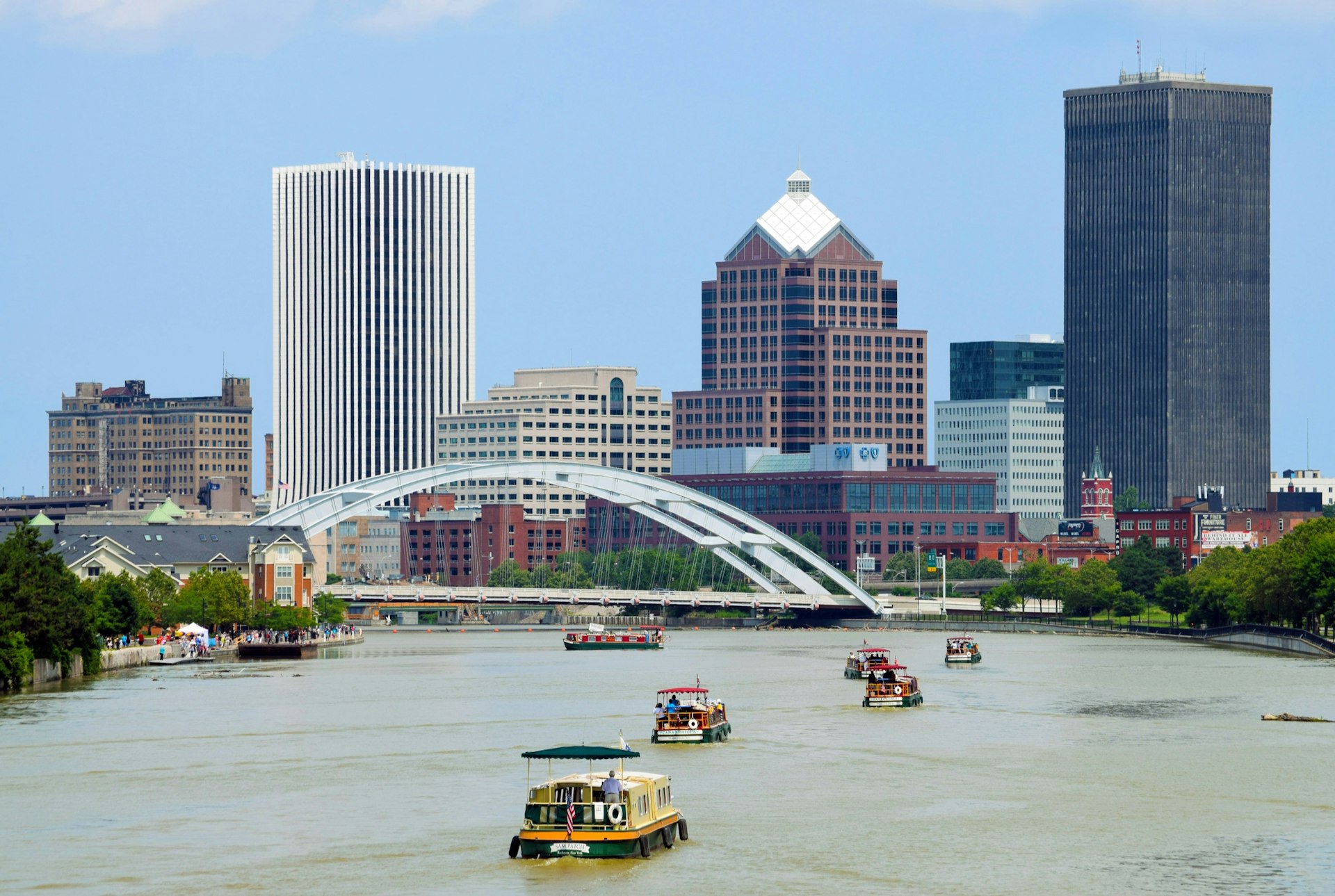 Several boats float down a wide river leading to a city skyline and arched bridge in Rochester, New York