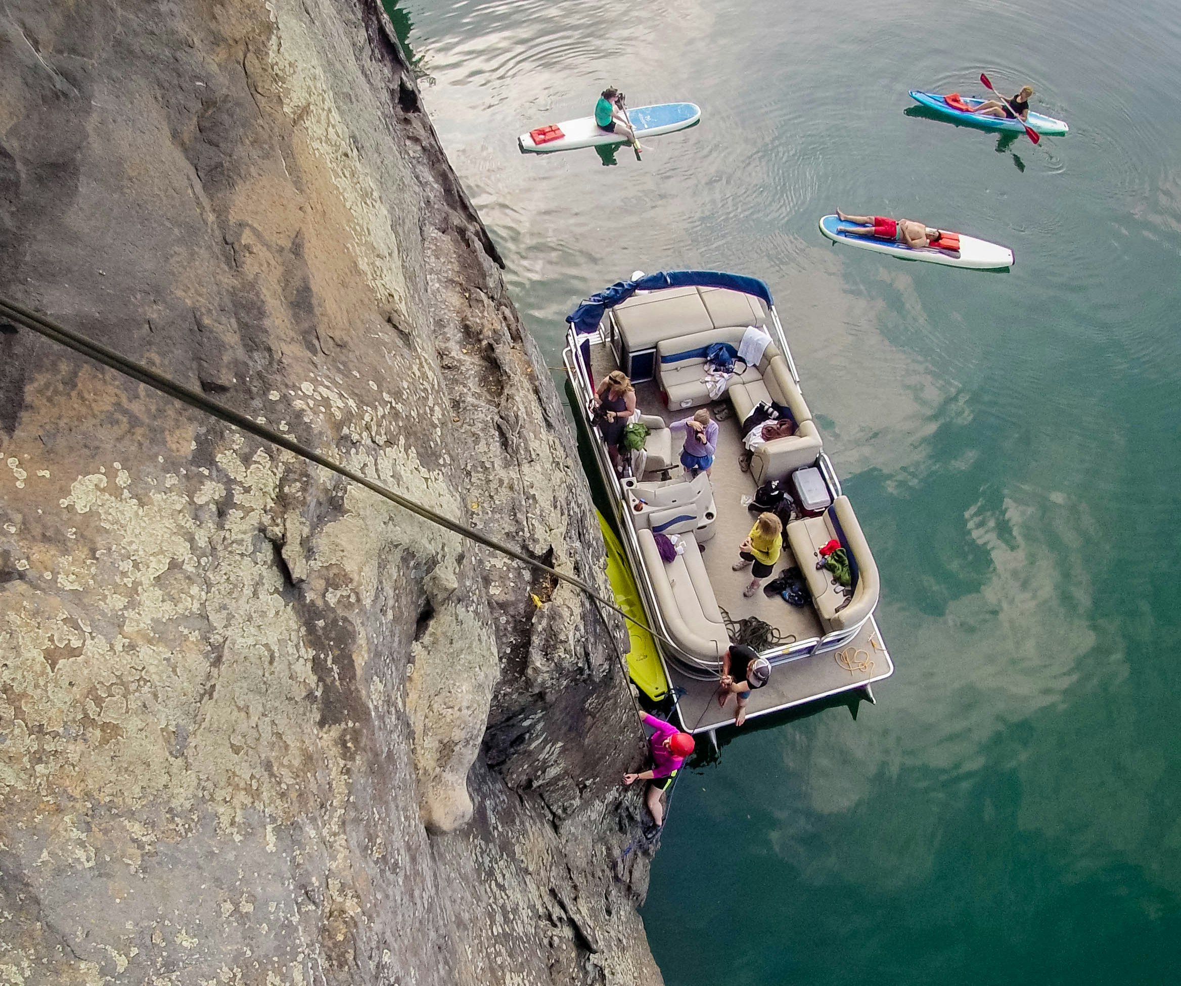 The camera is looking down on a boat which is next to a cliff face. A harness is hanging from the cliff, and a person climbing up the cliff face from the boat.