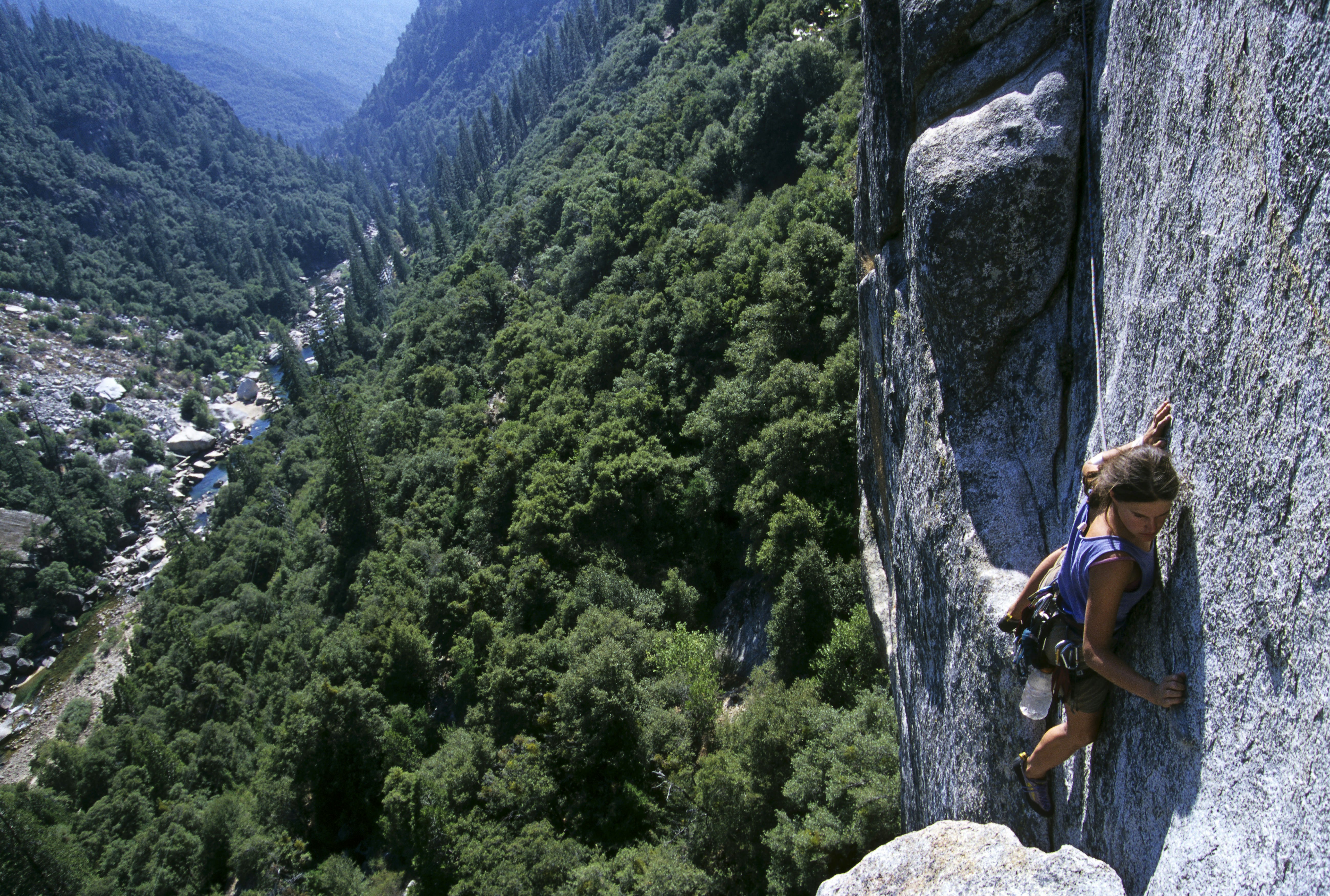 A rock climber clings to the cliff face while tackling an ascent in Yosemite National Park. Below her, a long way down, trees and a river are visible.
