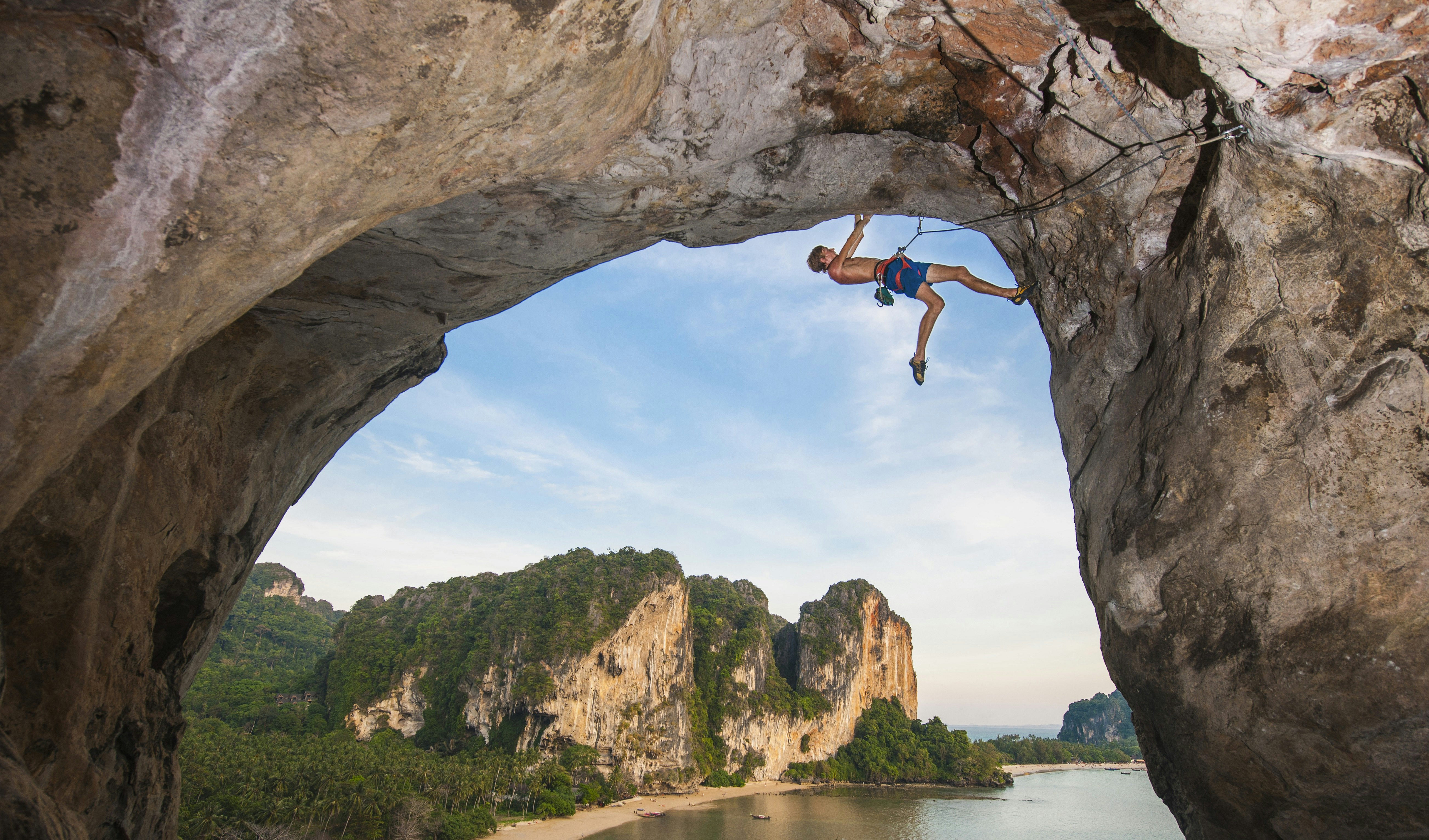 A climber dangles from an arching rock formation in Krabi province in Thailand. In the background large sea cliffs are visible, plus a strip of golden white sand.