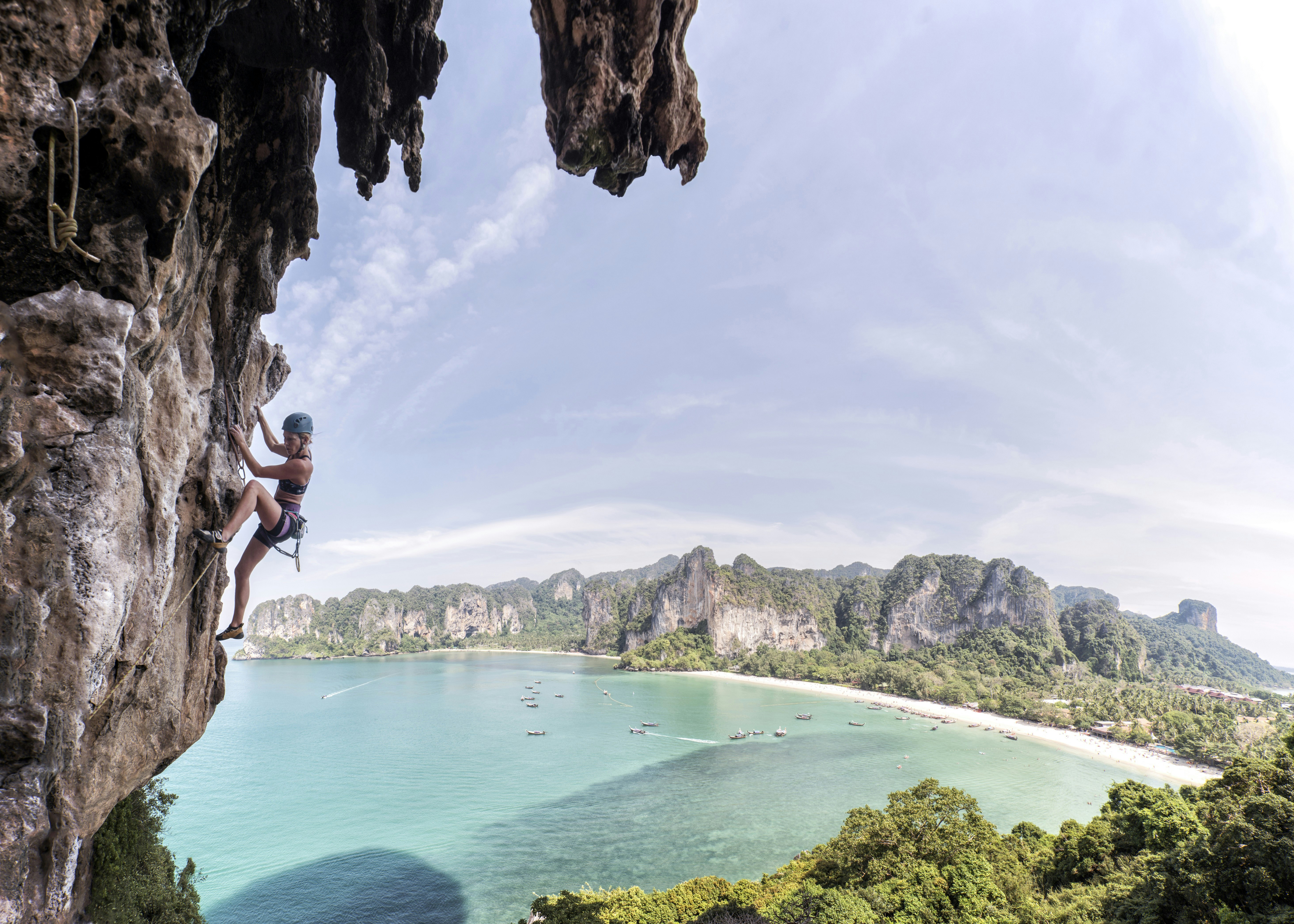 A rock climber scales a sheer rock face in Krabi, Thailand. In the background is a sweeping view of a white-sand beach and turquoise sea.