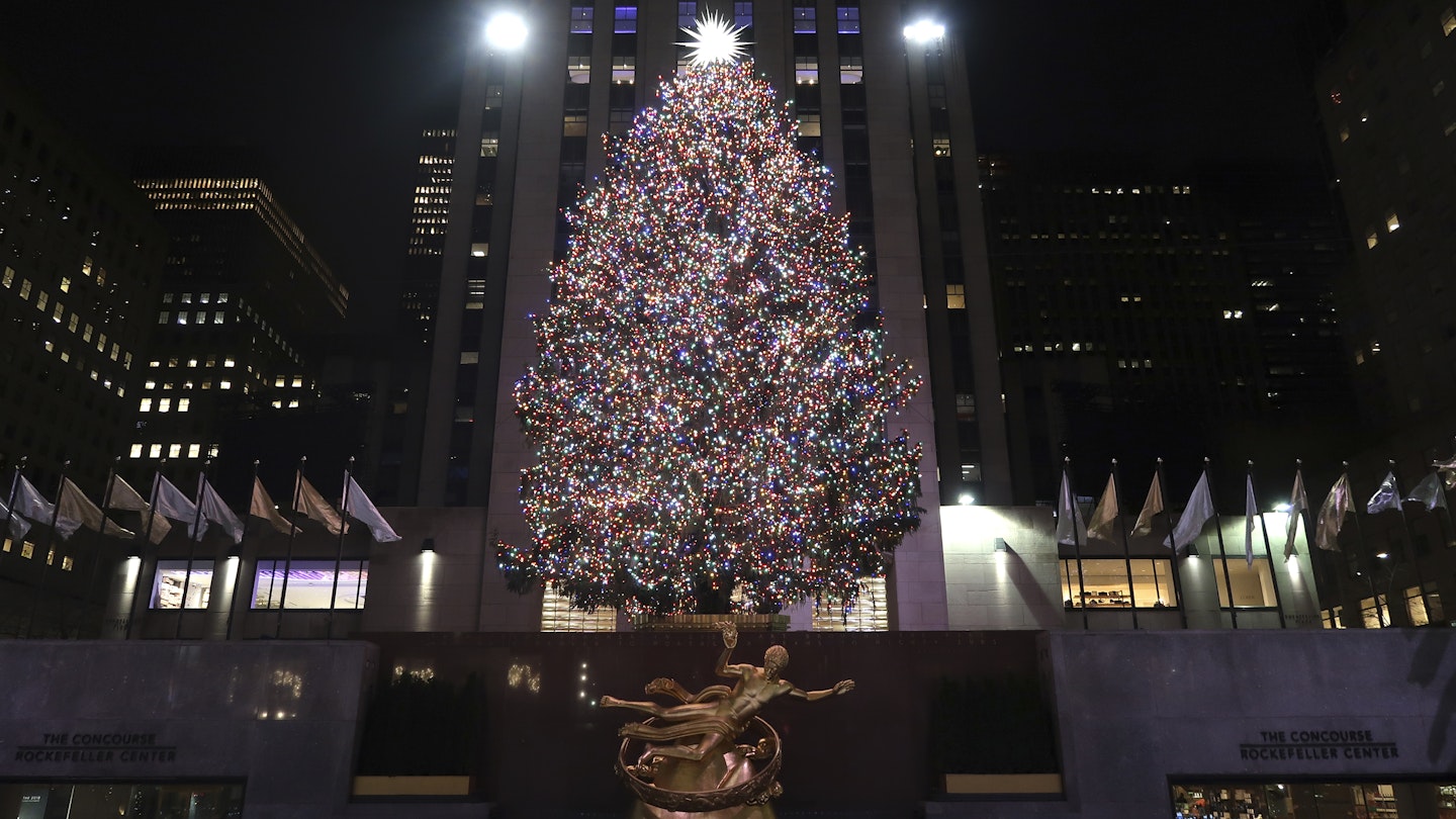 The Rockefeller Center Christmas tree shines brightly in front of the golden Prometheus statue at Rockefeller Center. There's a large bright start at the top of the tree.
