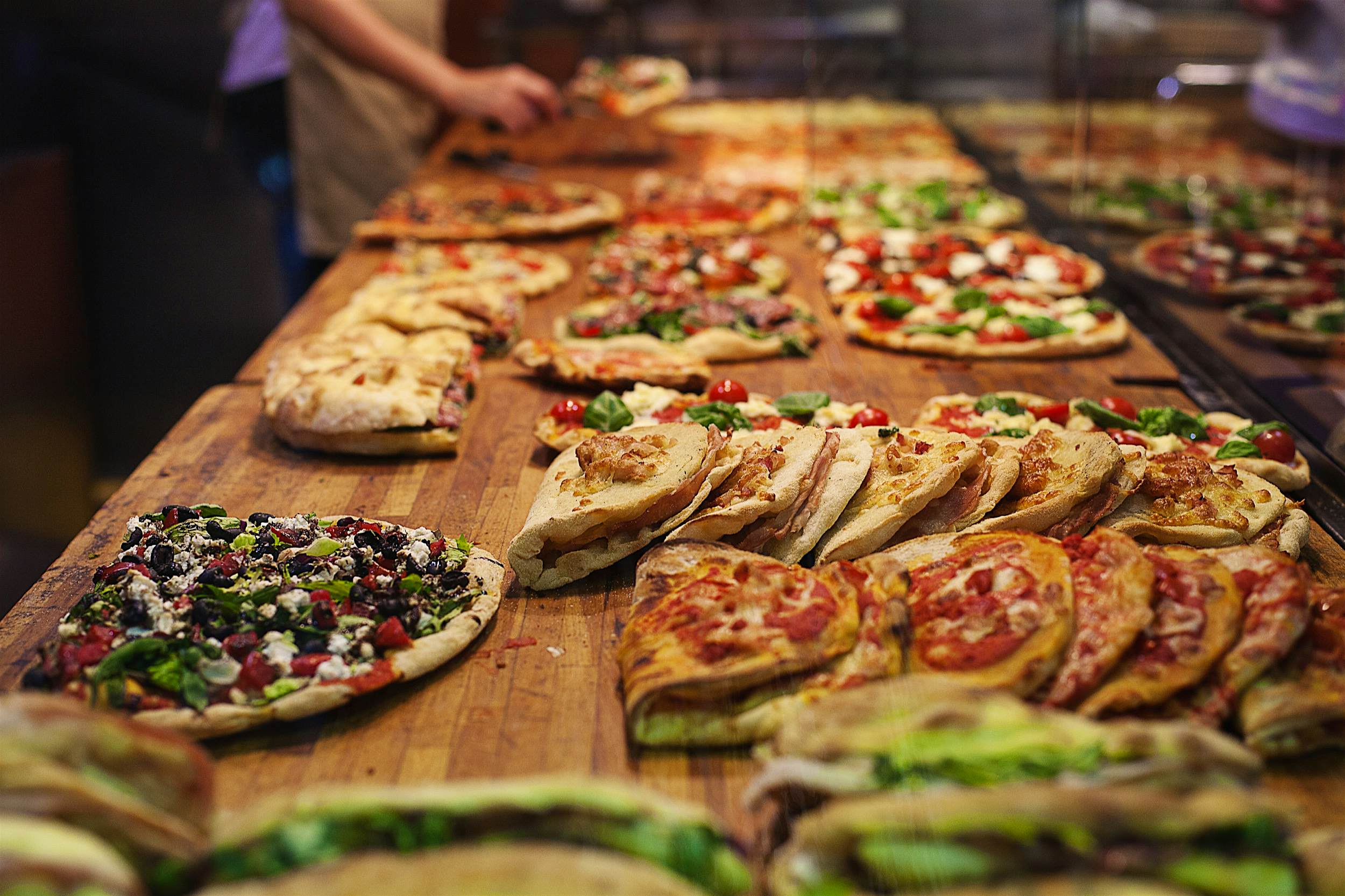 Roman Pizza.jpg A row of different styles of pizza sit behind a glass window on wooden boards.