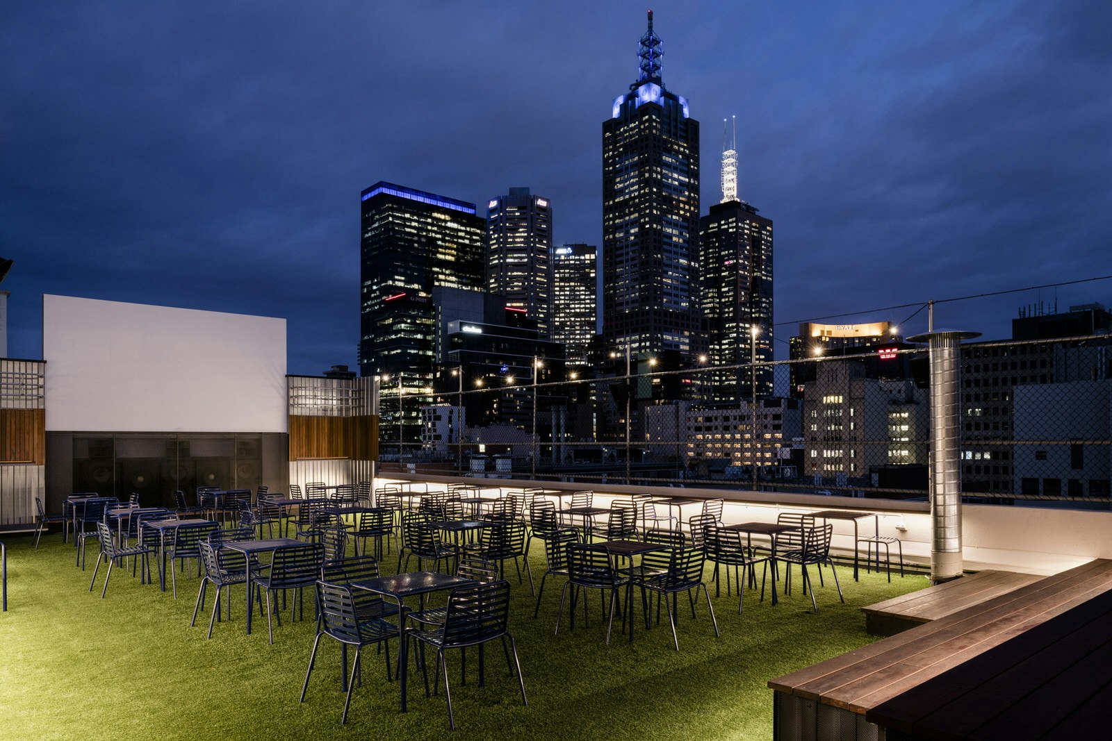 A nighttime shot of rooftop bar. There is astroturf on the floor, and tables and chairs are organised neatly in front of an outdoor cinema screen. Skyscrapers can be seen beyond the glass wall of the bar.
