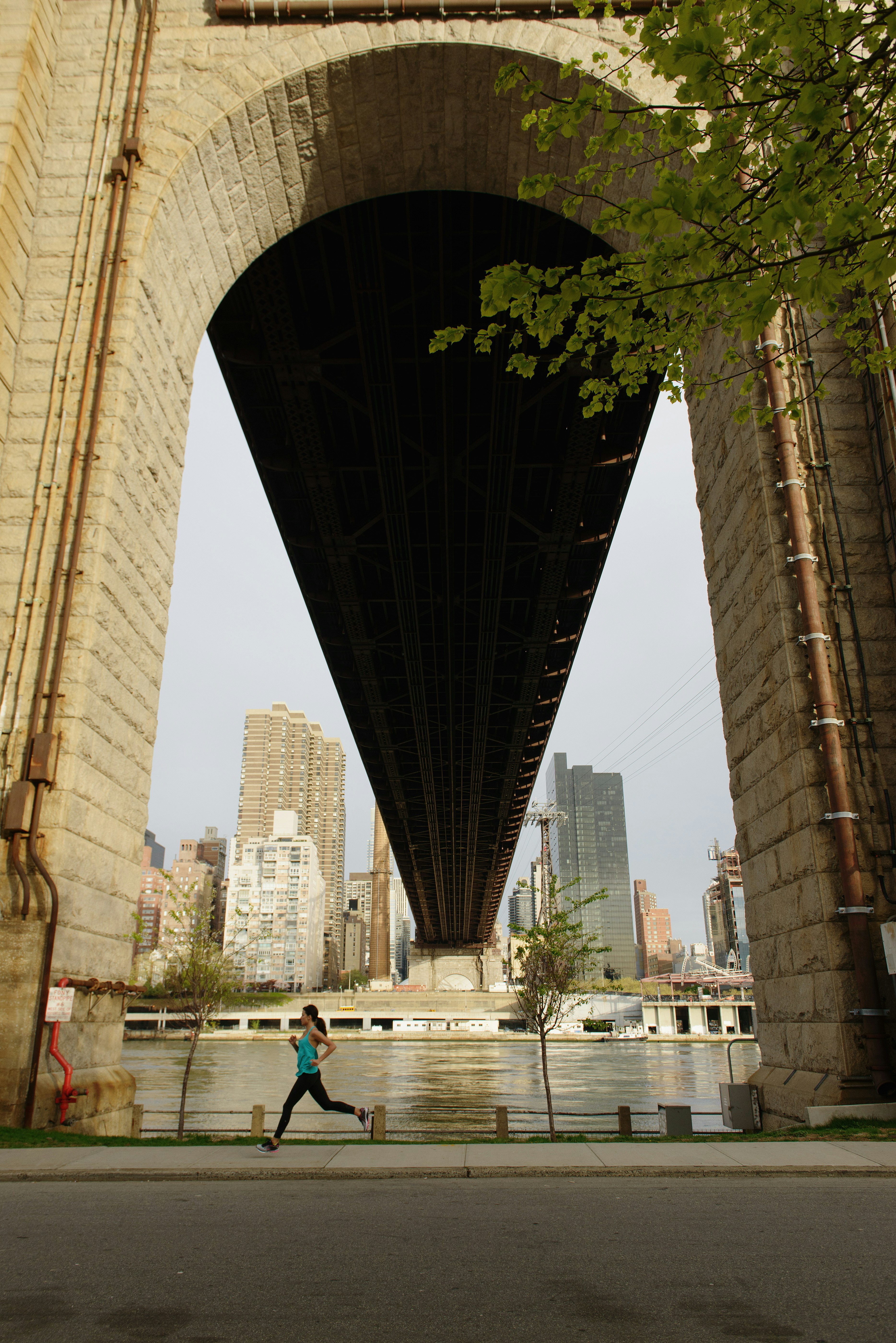 A woman is running on the sidewalk under one of the arches of the Queensboro Bridge on Roosevelt Island, New York. Across the river, skyscrapers and large buildings can be seen.
