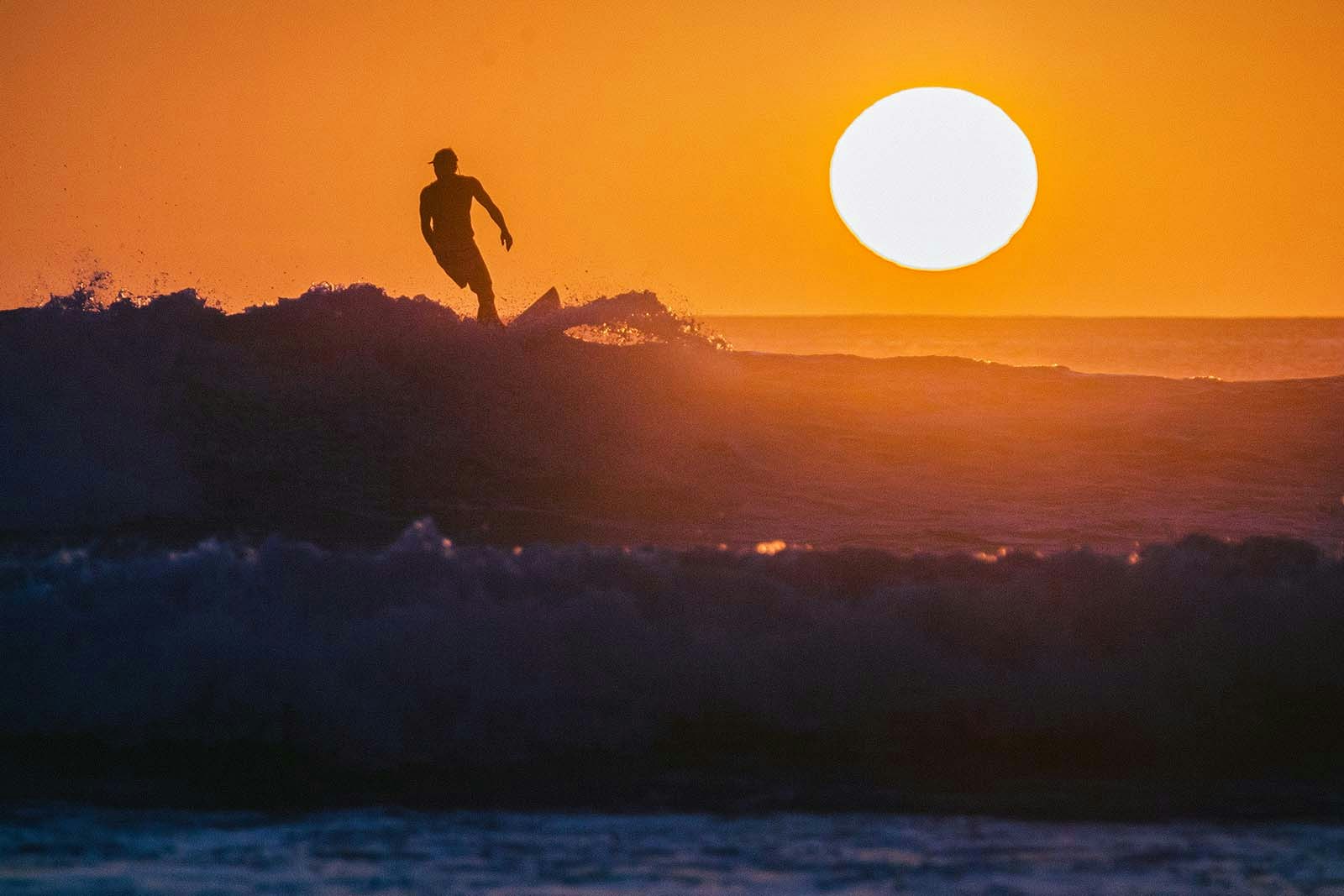 A surfer stands on his board at the crest of a wave; they are backlit by a setting orange sun. East Nusa Tenggara, Indonesia.