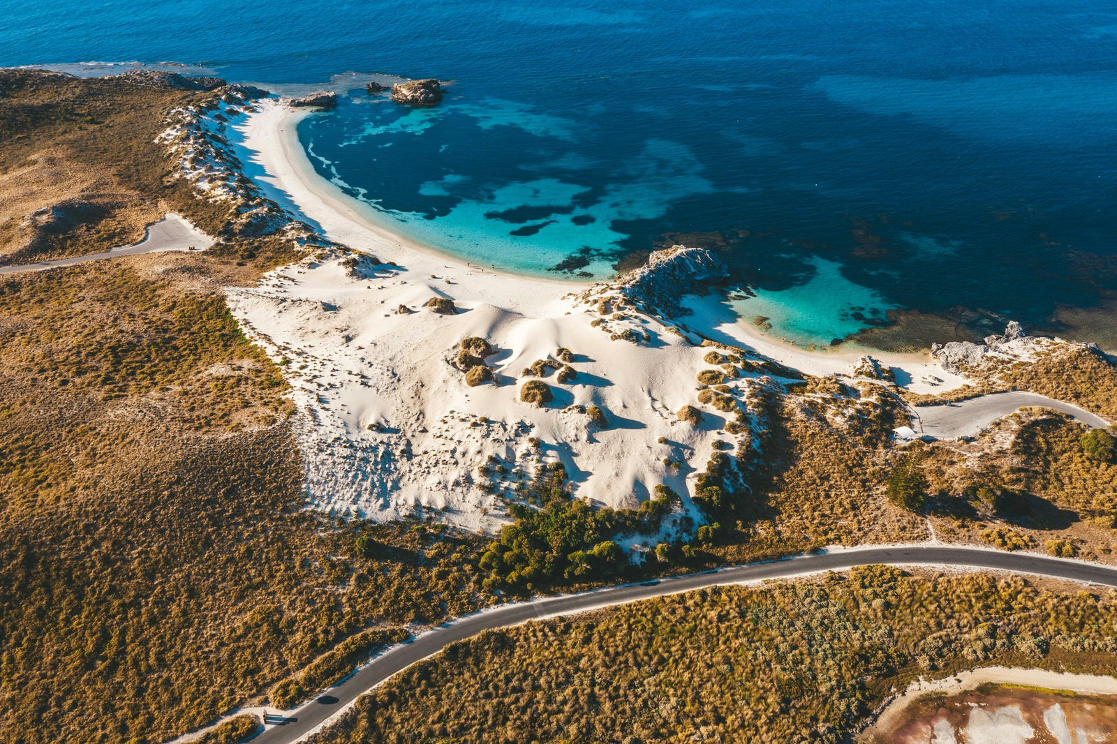 An aerial shot of Rottnest Island's coastline. Clear blue waters gently lap a white sandy beach that gives way to dunes and dense shrubbery