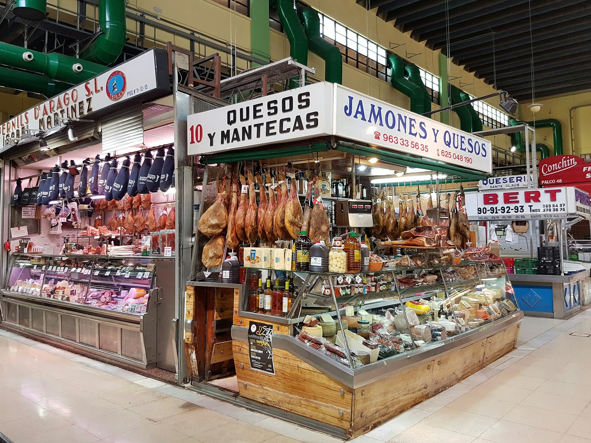 Stalls inside a brightly lit covered market. The closest stall has many cured hams hanging up on display, as well as an array of different cheeses in a glass cabinet.