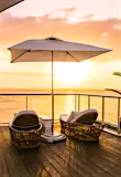 A collection of rattan lounge chairs, a large umbrella and a large potted plant face the ocean as the sun sets on the sky deck area at the S Hotel in Jamaica.