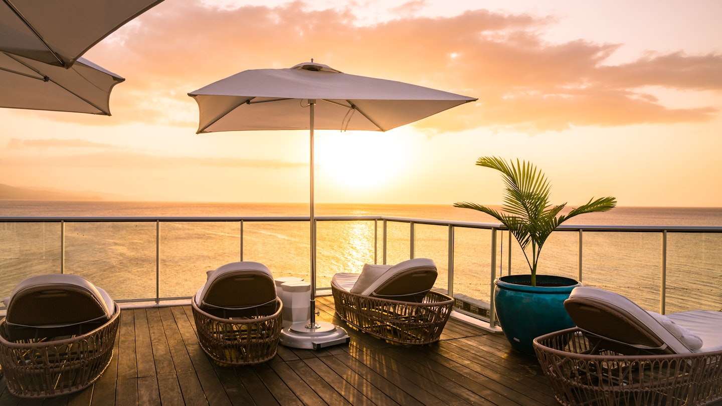 A collection of rattan lounge chairs, a large umbrella and a large potted plant face the ocean as the sun sets on the sky deck area at the S Hotel in Jamaica. 