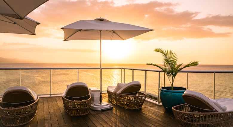 A collection of rattan lounge chairs, a large umbrella and a large potted plant face the ocean as the sun sets on the sky deck area at the S Hotel in Jamaica. 