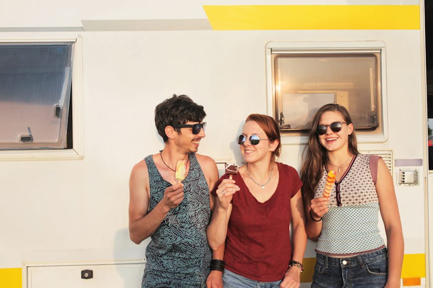 Friends eat ice cream cones as they lean up against a camper. They have sunglasses on and tank tops. California Ice Cream