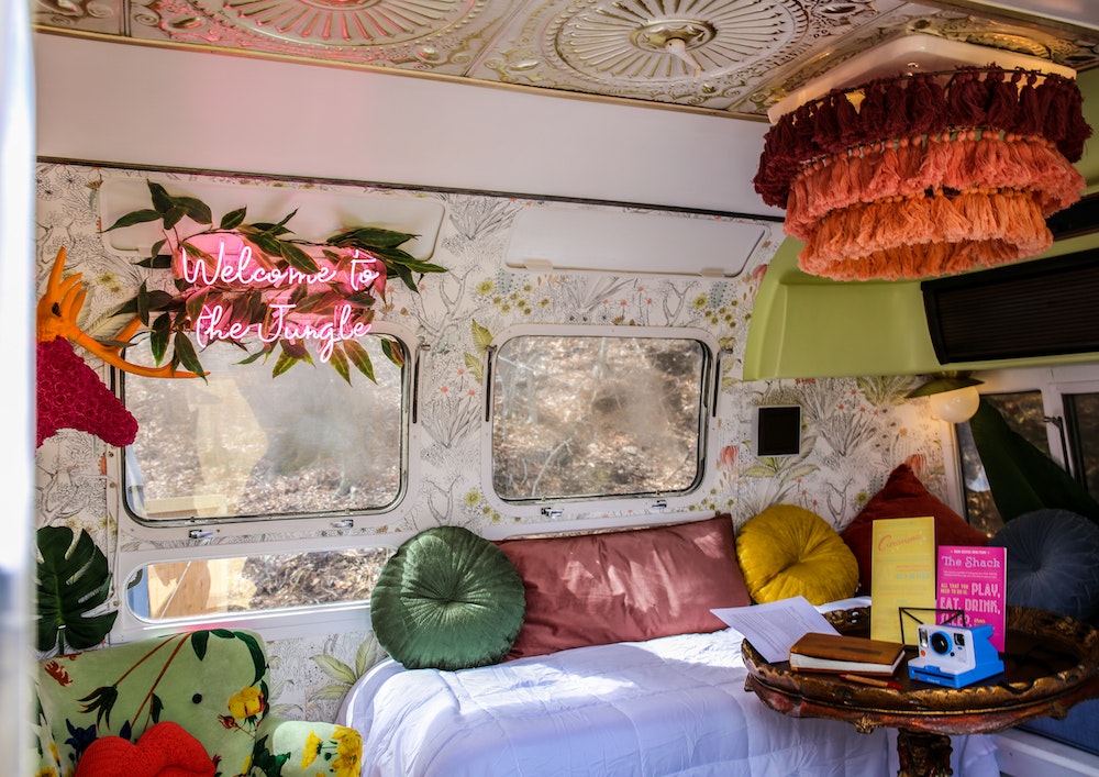 The interior of one of the Airstream rooms at Hotel Caravana is wallpapered in a pen-and-ink style design of flowers and plants with pops of orange and green. A white seating bench has round velvet pillows in green, pink, and yellow. A pink neon sign over one of hte windows reads "Welcome to the Jungle" in cursive. A crocheted red deer head with orange antlers is on the left next to a monsterra leaf. A gilt table holds a blue Polaroid camera. A lampshade overhead is made of maroon and orange tassels.
